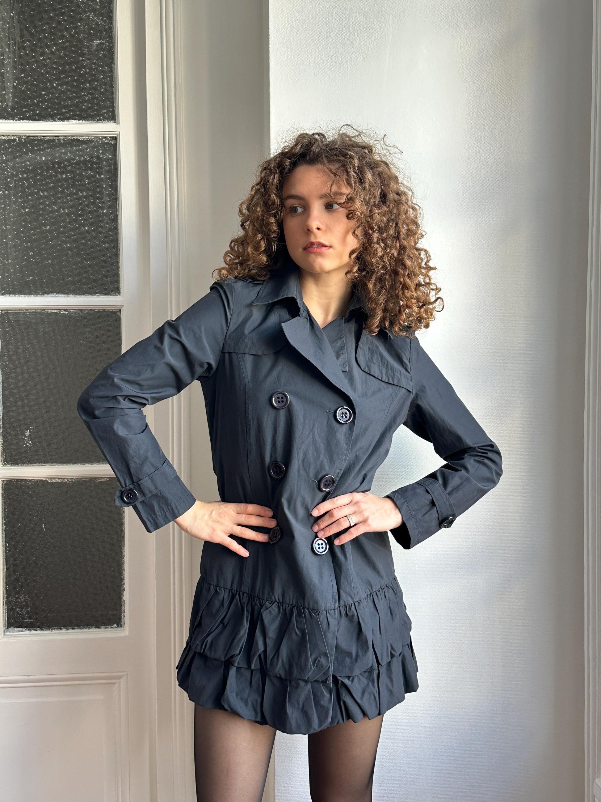 A woman with curly hair wearing a gray coat and skirt is posing in a relaxed manner.