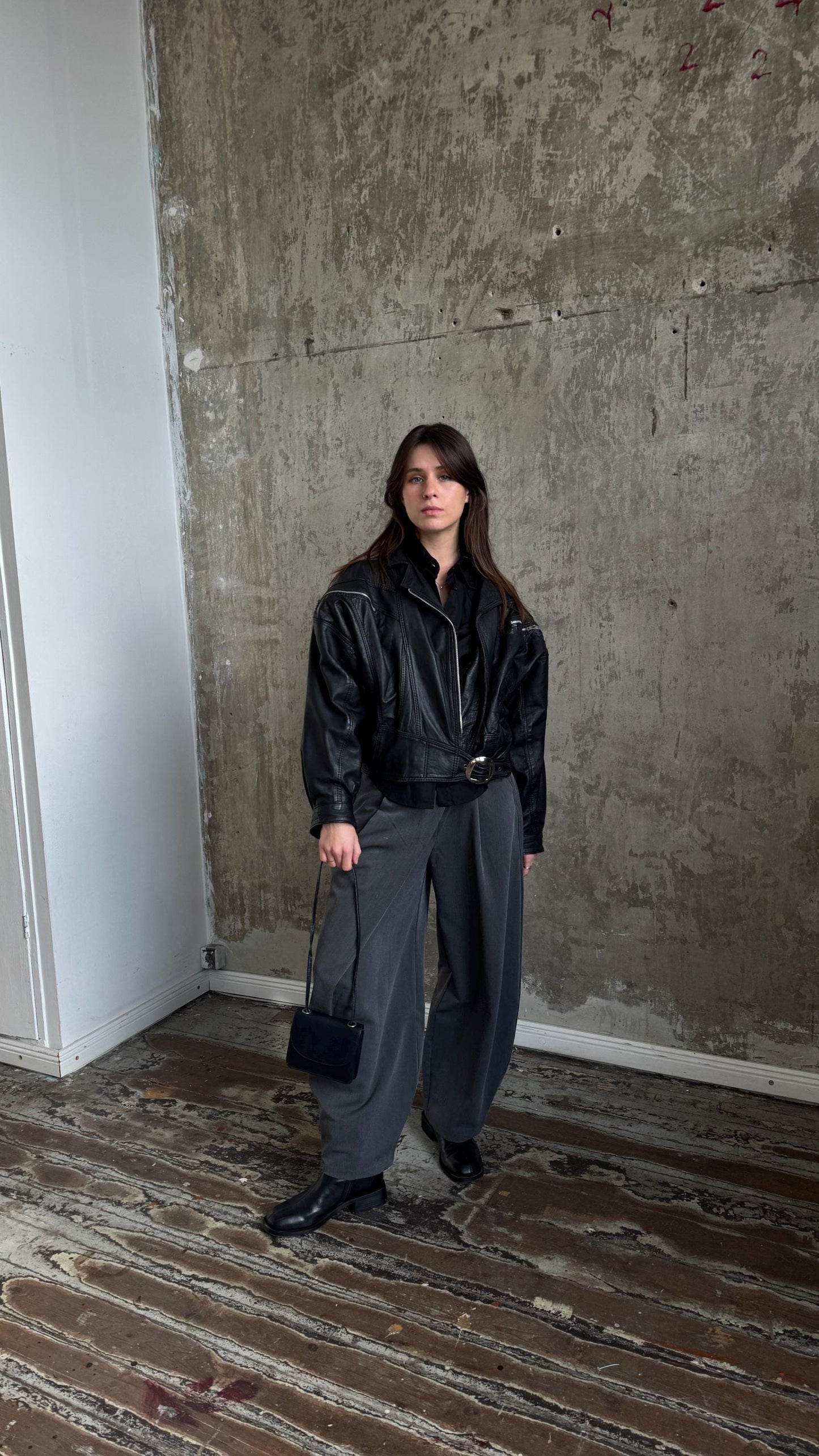 A woman wearing a black leather jacket and gray pants stands in a room with a concrete wall.