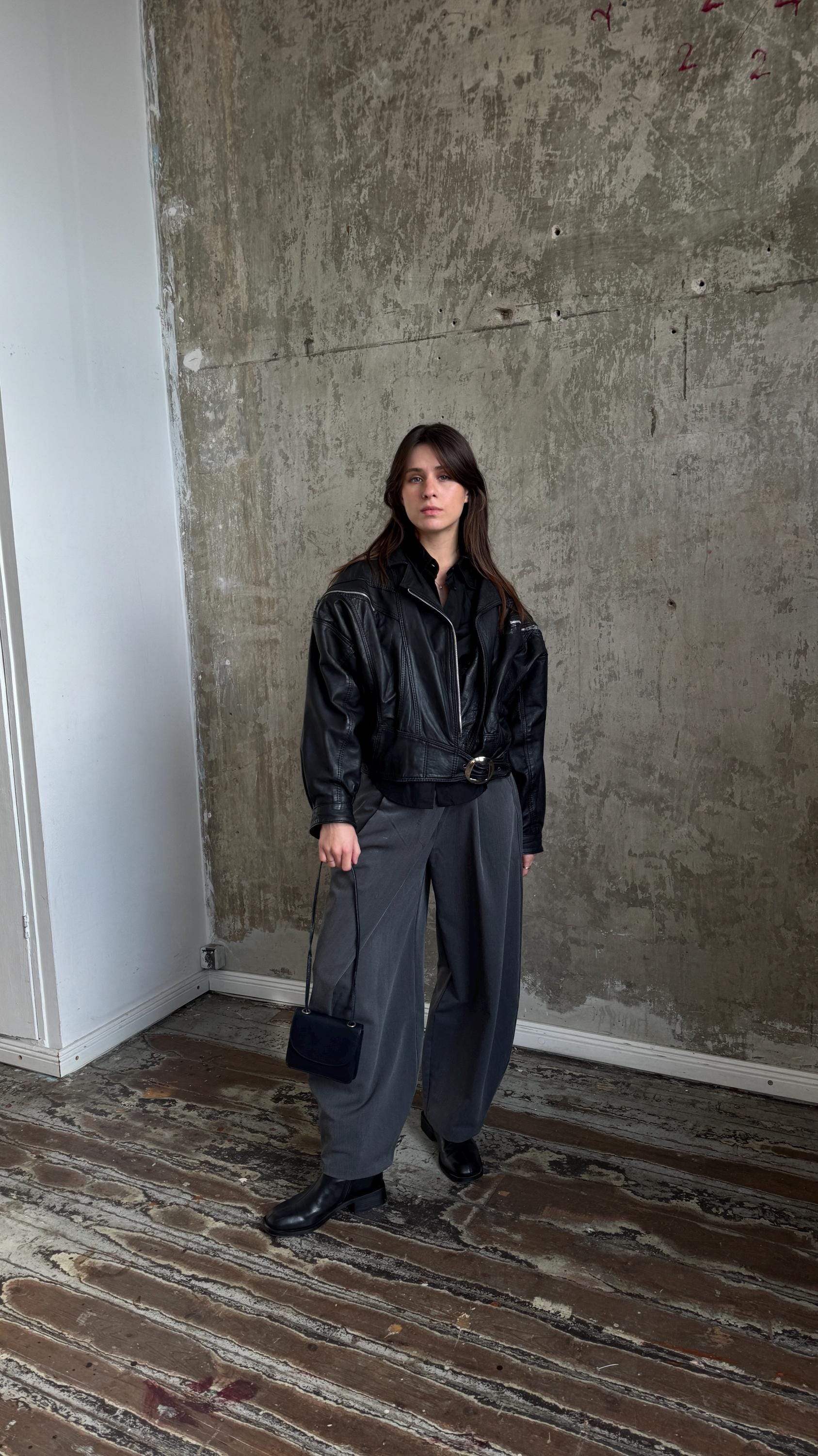 A woman wearing a black leather jacket and gray pants stands in a room with a concrete wall.