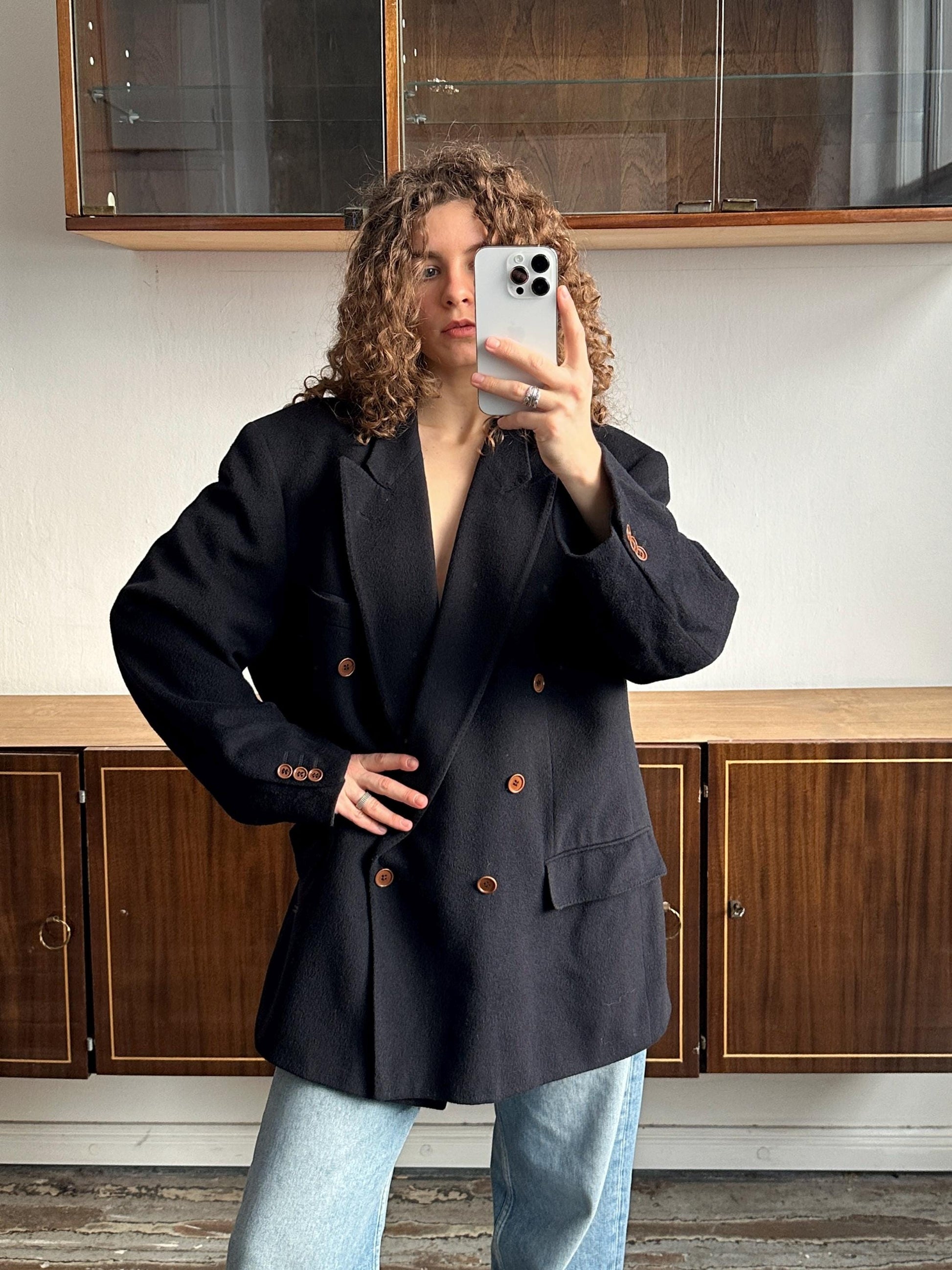 A person with curly hair is taking a selfie with a white phone, wearing a black blazer and jeans.