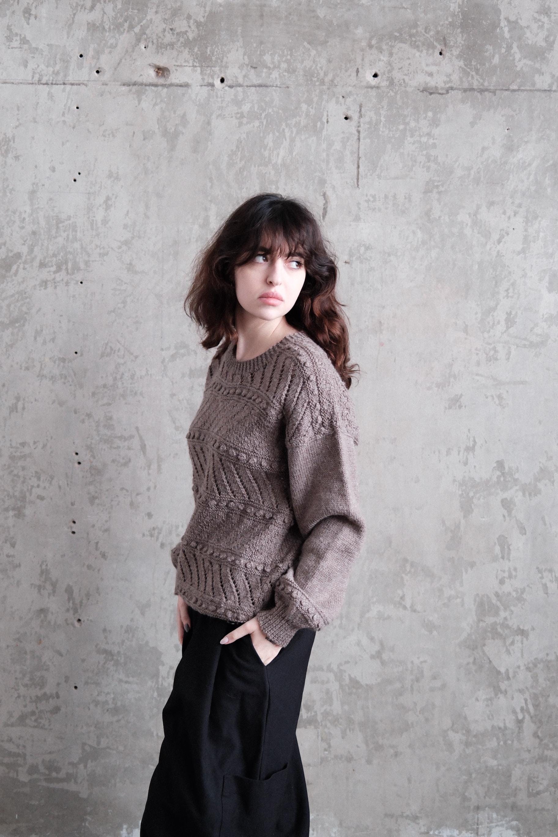 A woman with long dark hair is standing against a concrete wall, wearing a gray sweater and black pants.