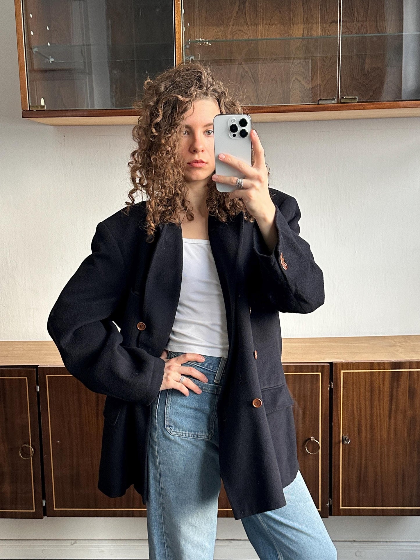 A woman with curly hair is taking a selfie with a smartphone, wearing a black blazer and jeans.
