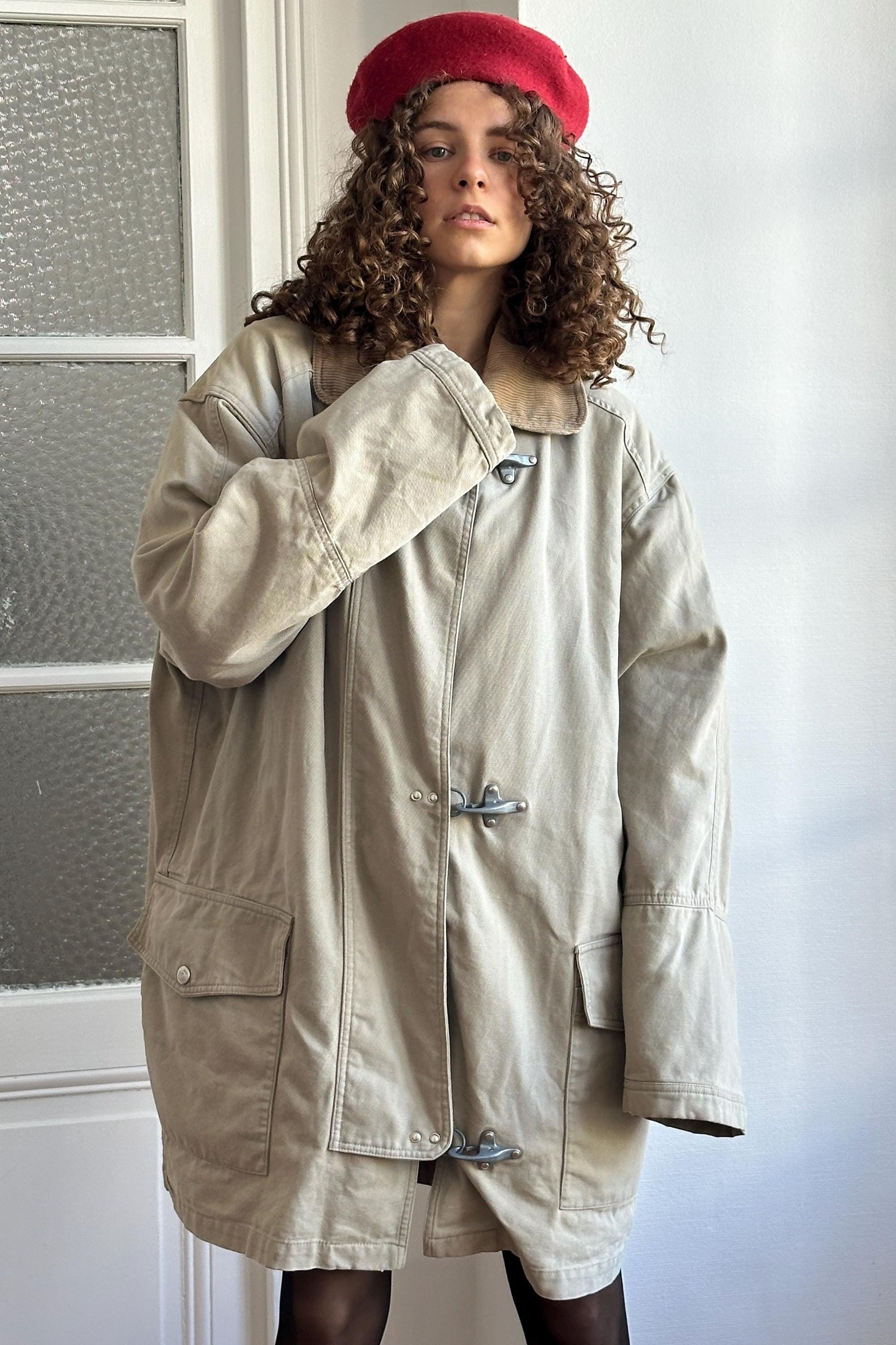 A woman with curly hair wearing a beige coat and a red hat stands in front of a white door.