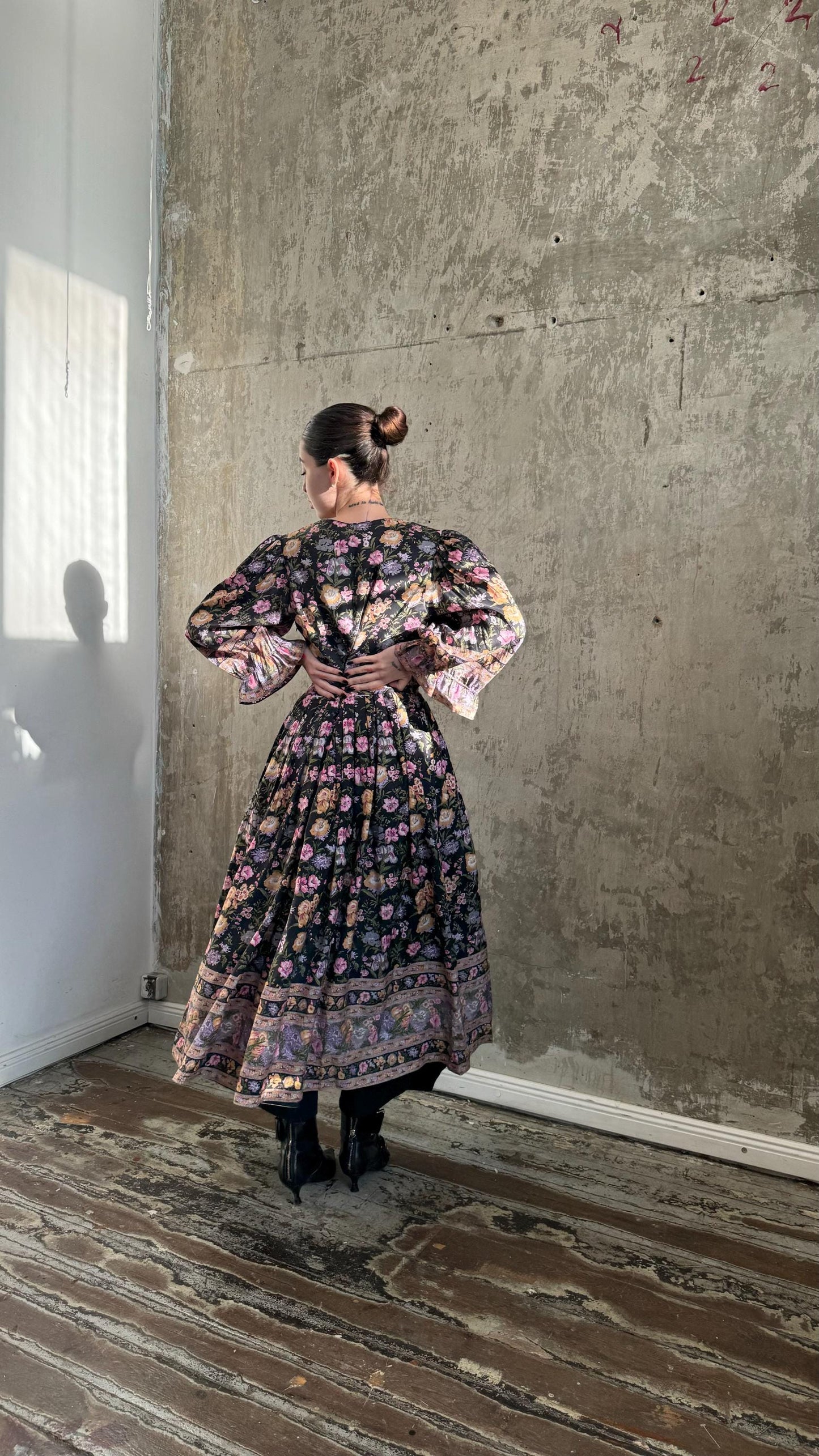 A woman wearing a long, floral-patterned dress stands in a room with a concrete wall and wooden floor.