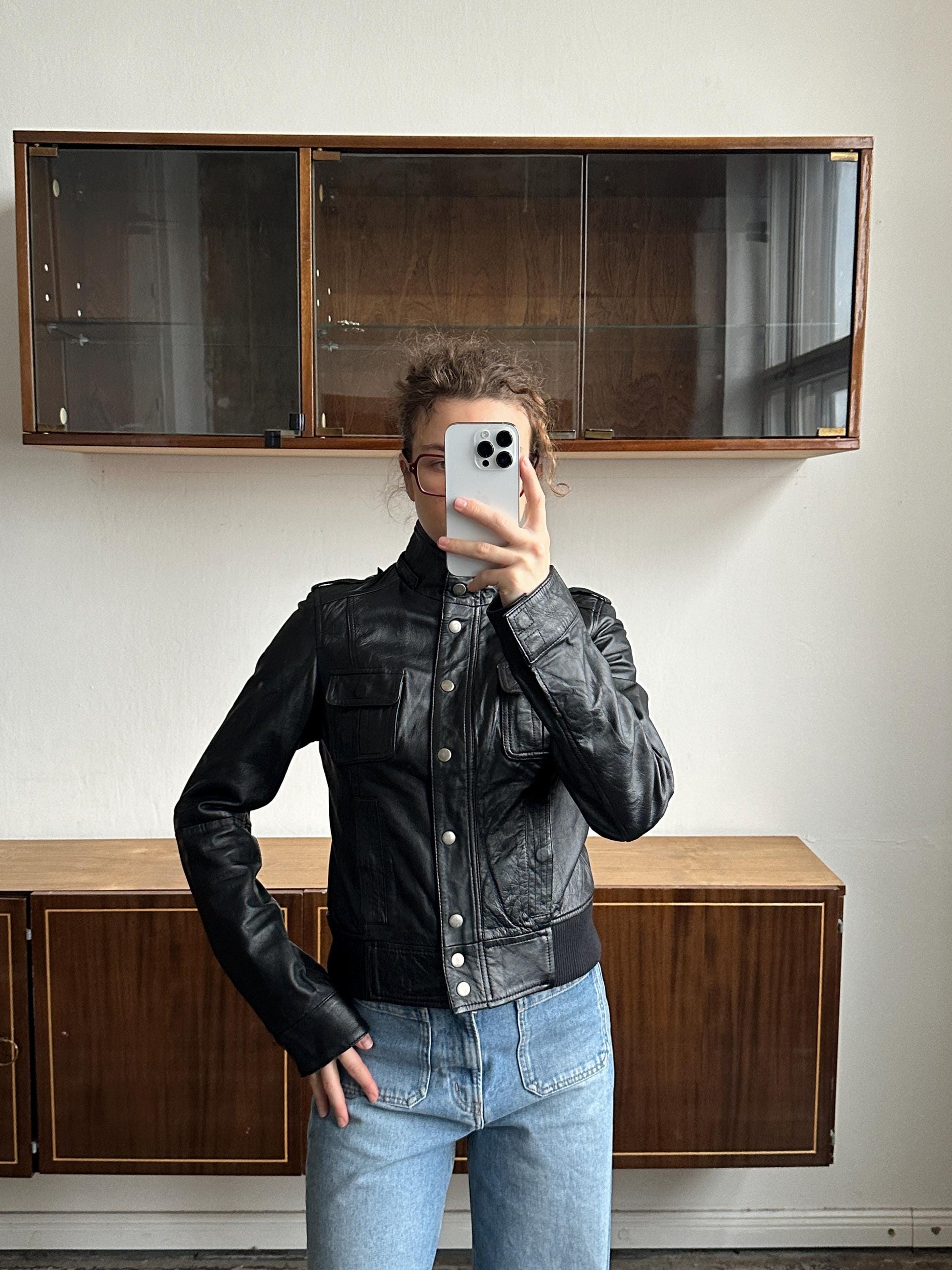 A person is taking a selfie with a smartphone, wearing a black leather jacket and jeans, in front of a wooden cabinet.
