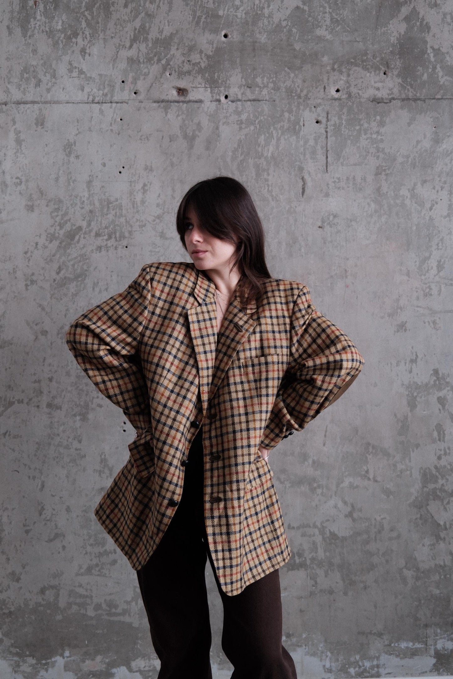 A person wearing a plaid jacket stands against a concrete wall, with their arms crossed.