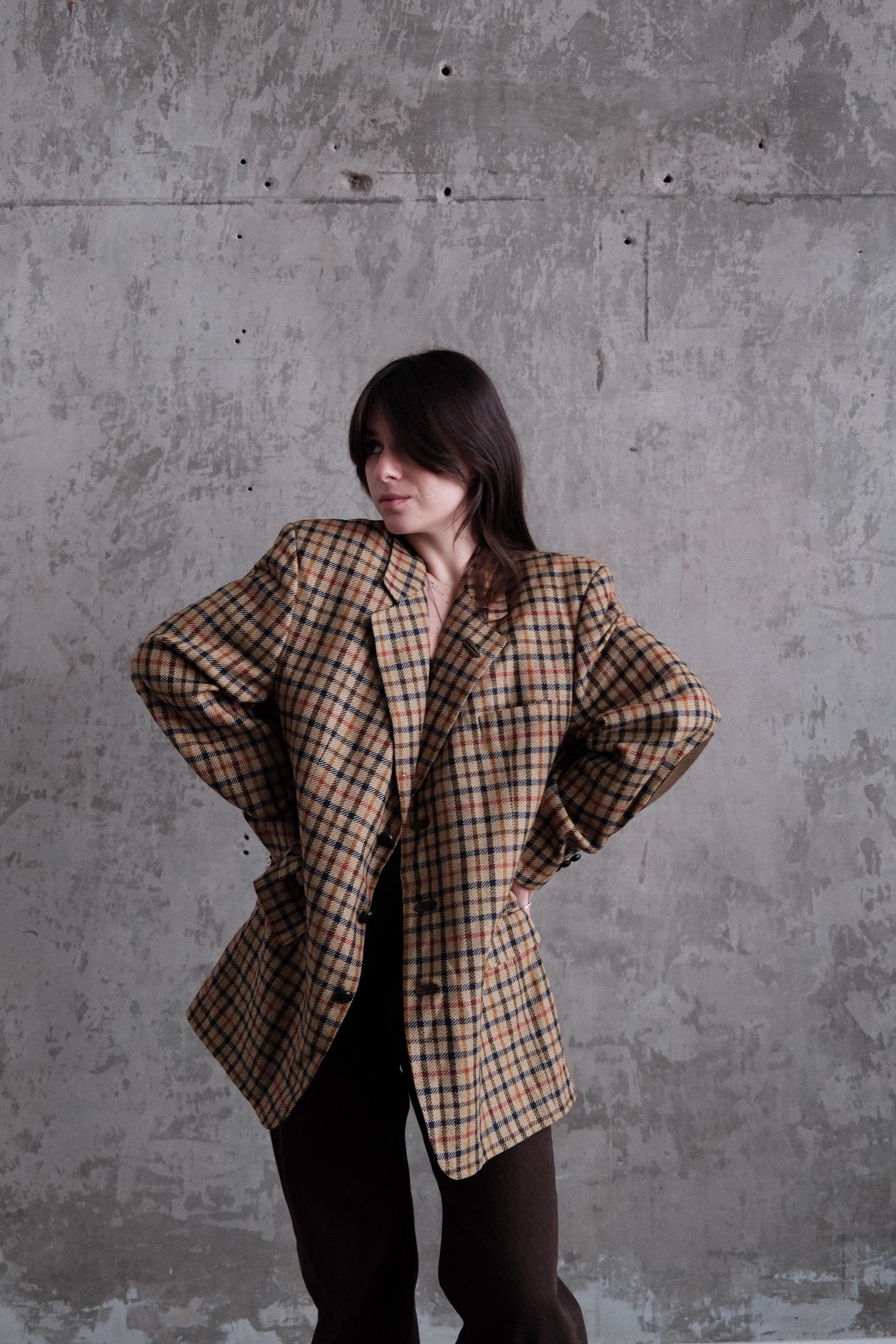 A person wearing a plaid jacket stands against a concrete wall, with their arms crossed.