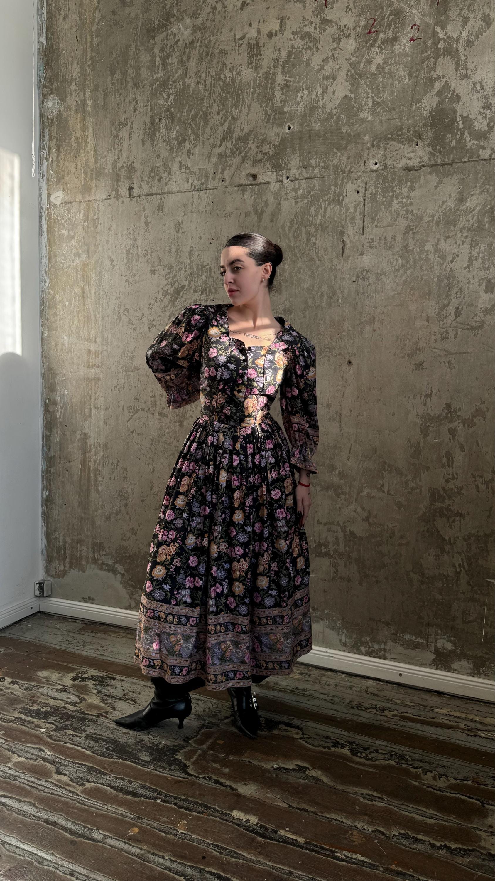 A woman wearing a long, floral-patterned dress stands in a room with a concrete wall.
