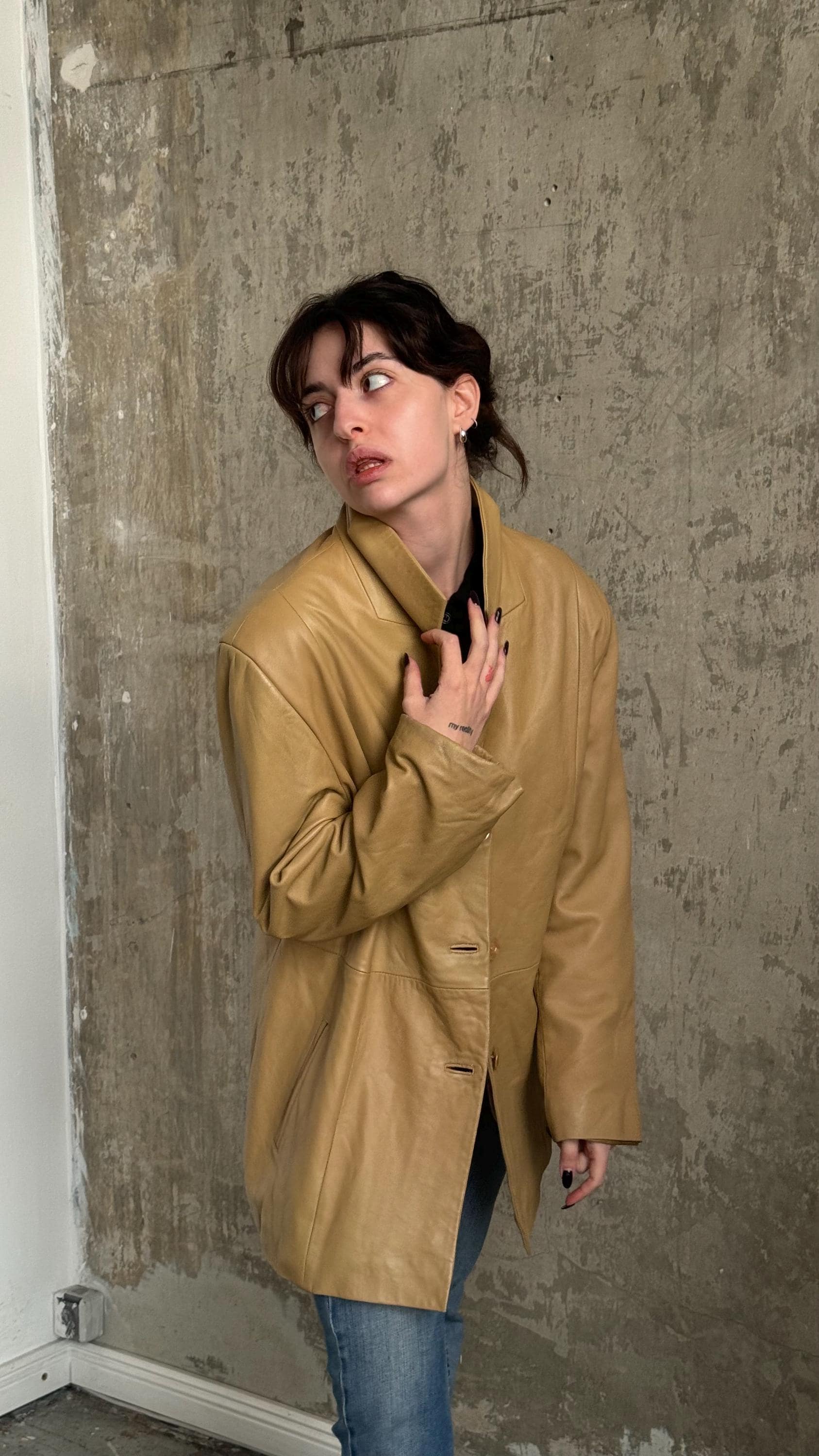 A woman wearing a tan leather jacket stands in front of a concrete wall, looking off to the side with a serious expression.