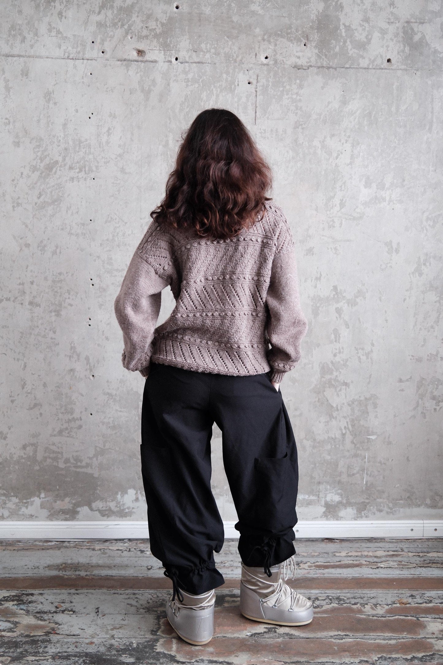 A person wearing a gray sweater and black pants stands in front of a concrete wall, with their back turned to the camera.