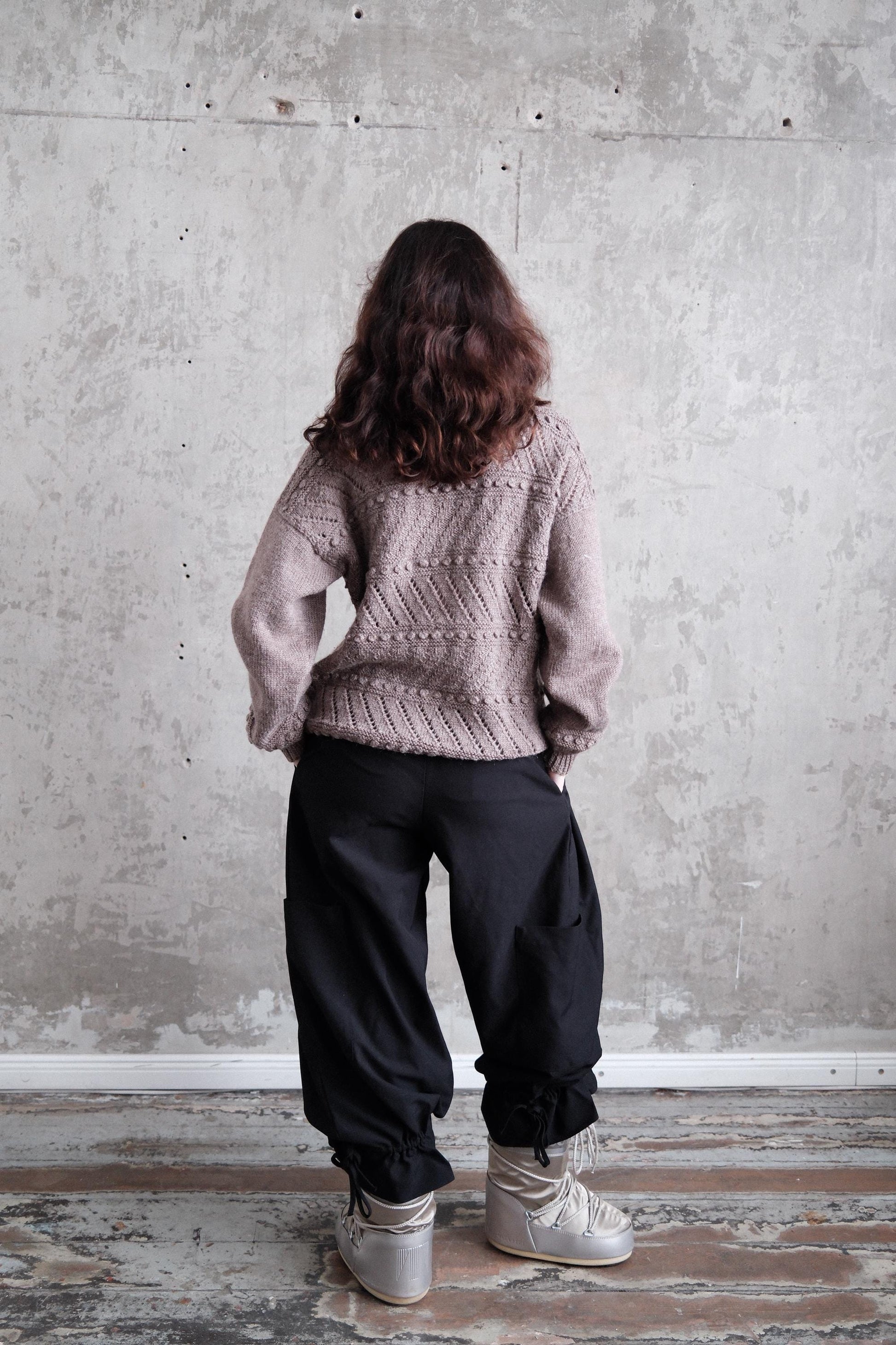 A person wearing a gray sweater and black pants stands in front of a concrete wall, with their back turned to the camera.