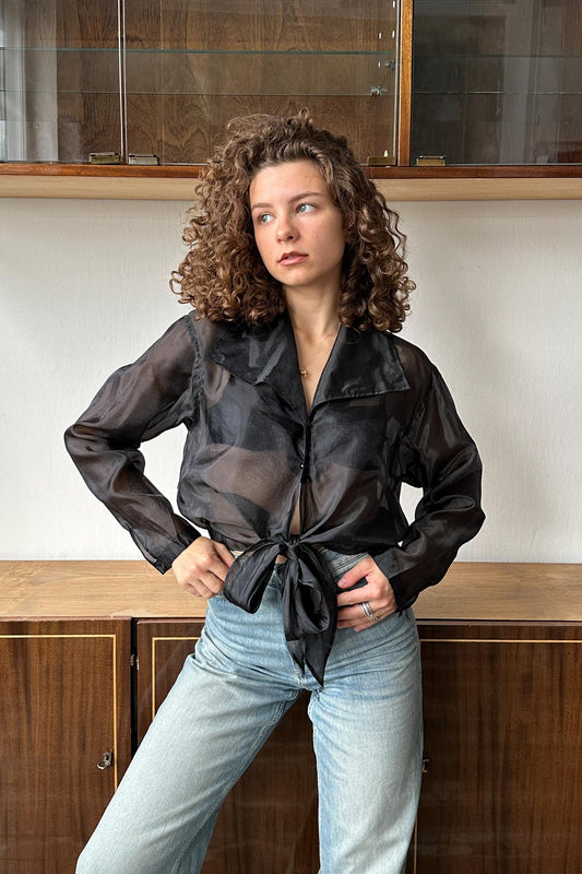 A woman with curly hair wearing a black blouse and light blue jeans stands in front of a wooden cabinet.