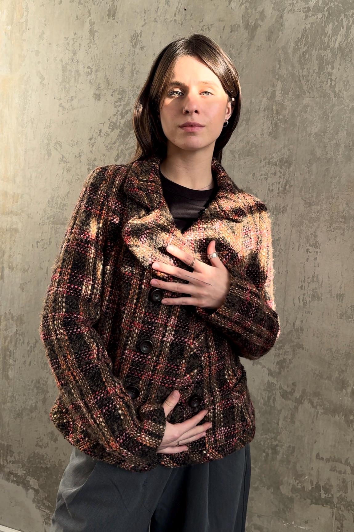 A woman wearing a brown and black plaid jacket stands in front of a concrete wall, her hands placed on her chest.