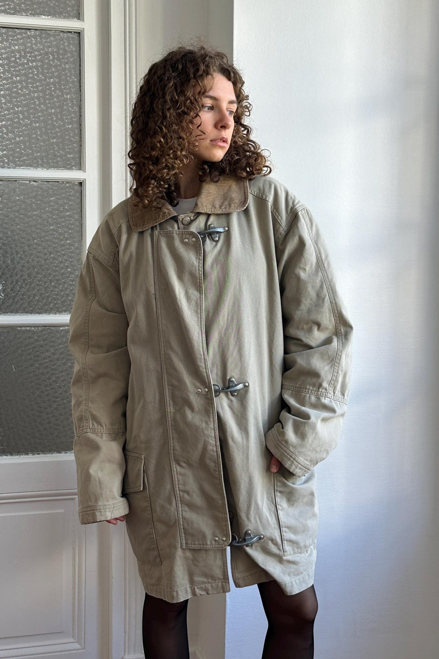 A woman with curly hair wearing a beige coat stands in front of a window, looking off to the side.