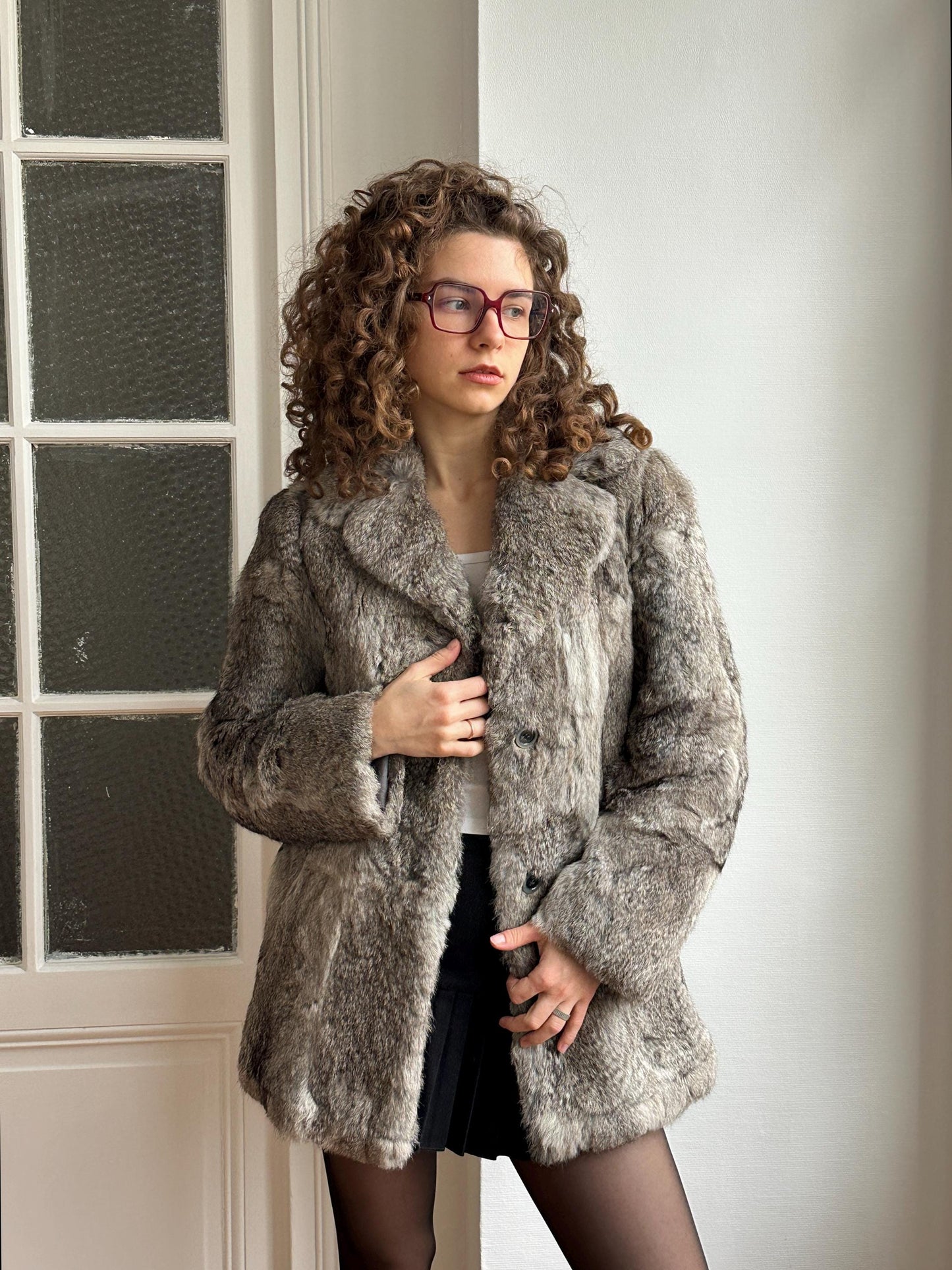 A woman with curly hair and glasses stands in front of a white door, wearing a gray fur coat and black skirt.