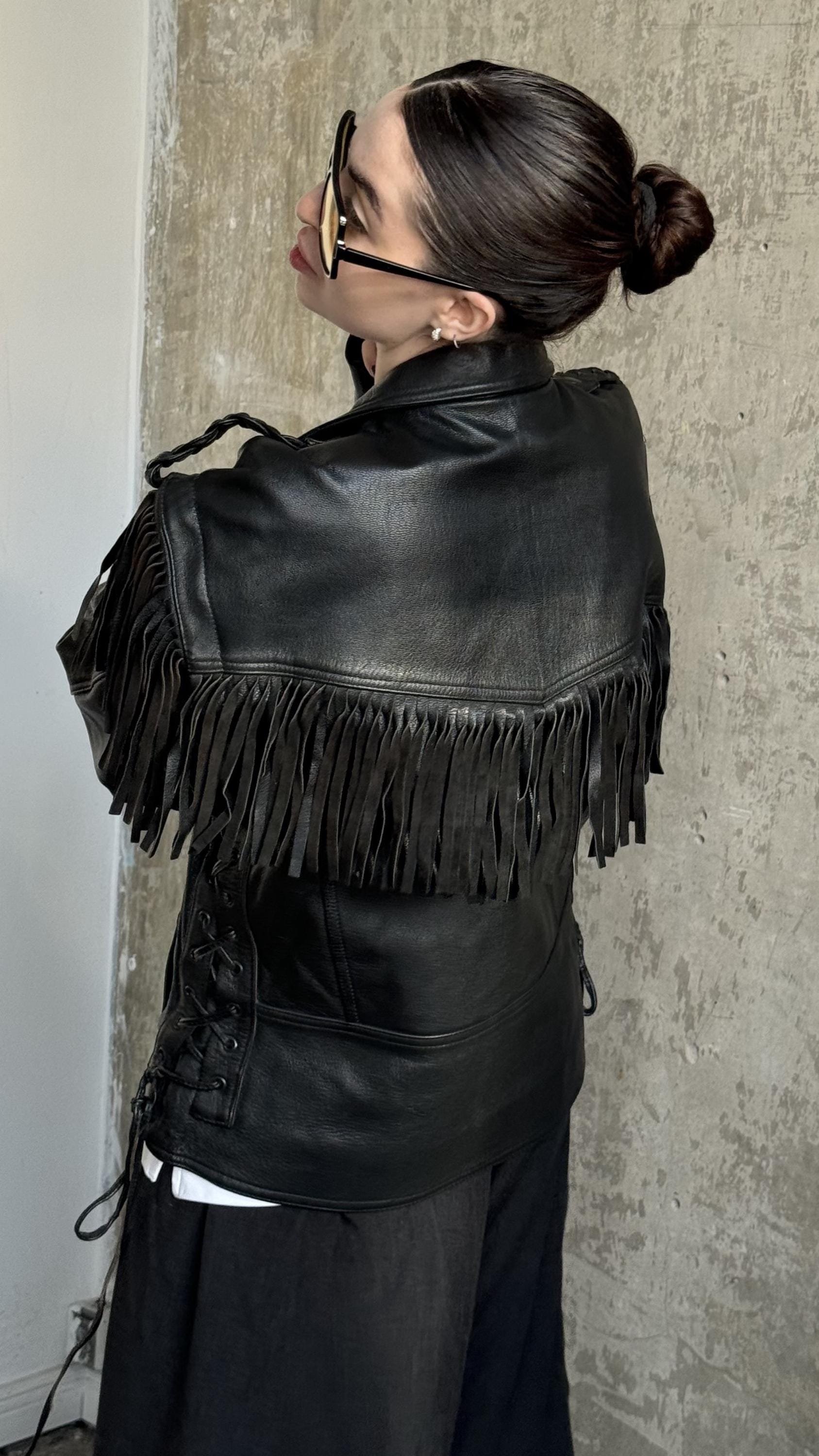 A woman wearing a black leather jacket with fringe, sunglasses, and a bun hairstyle stands against a concrete wall.