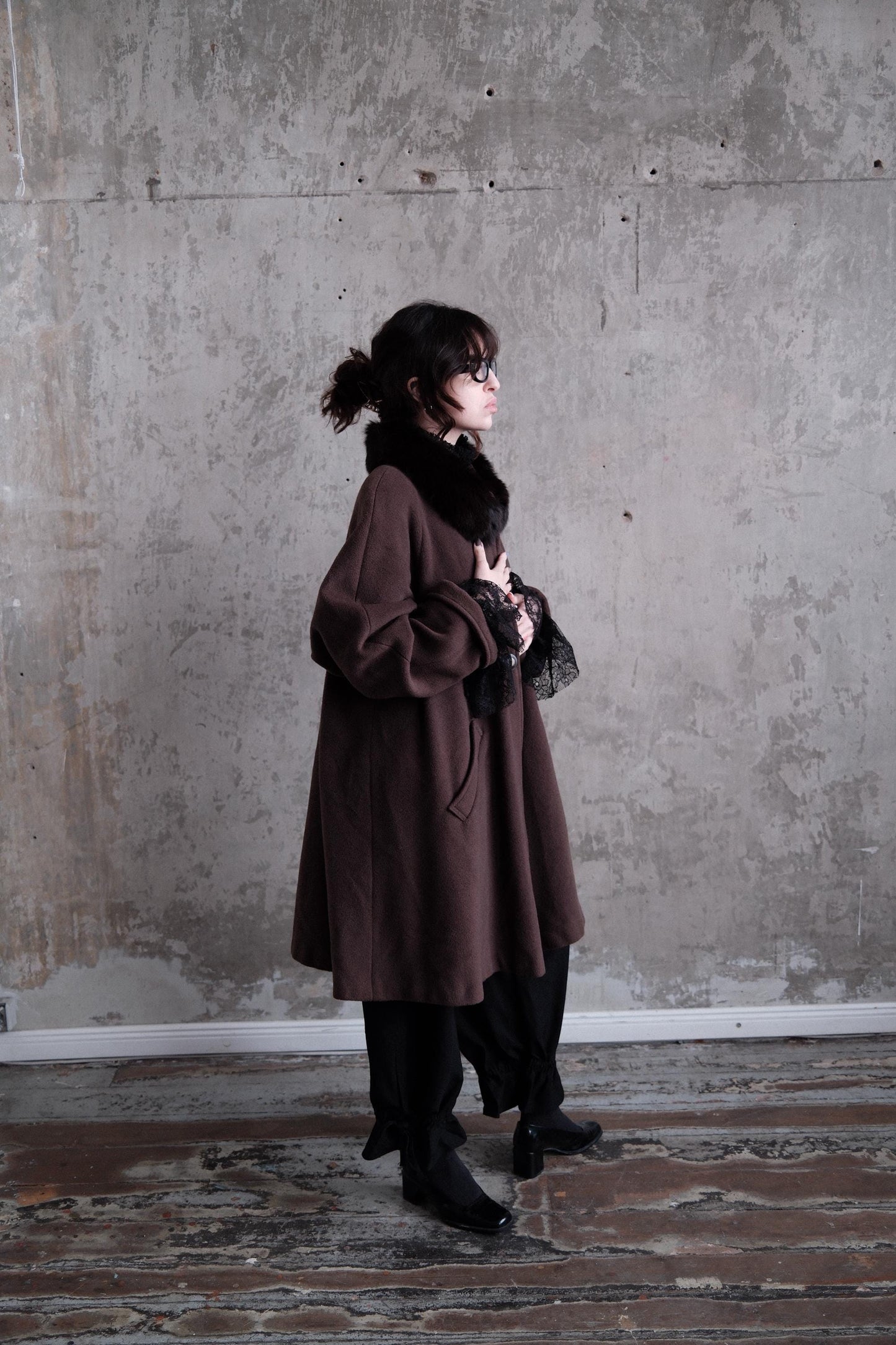 A woman in a brown coat stands in a room with a concrete wall, her gaze directed off to the side.