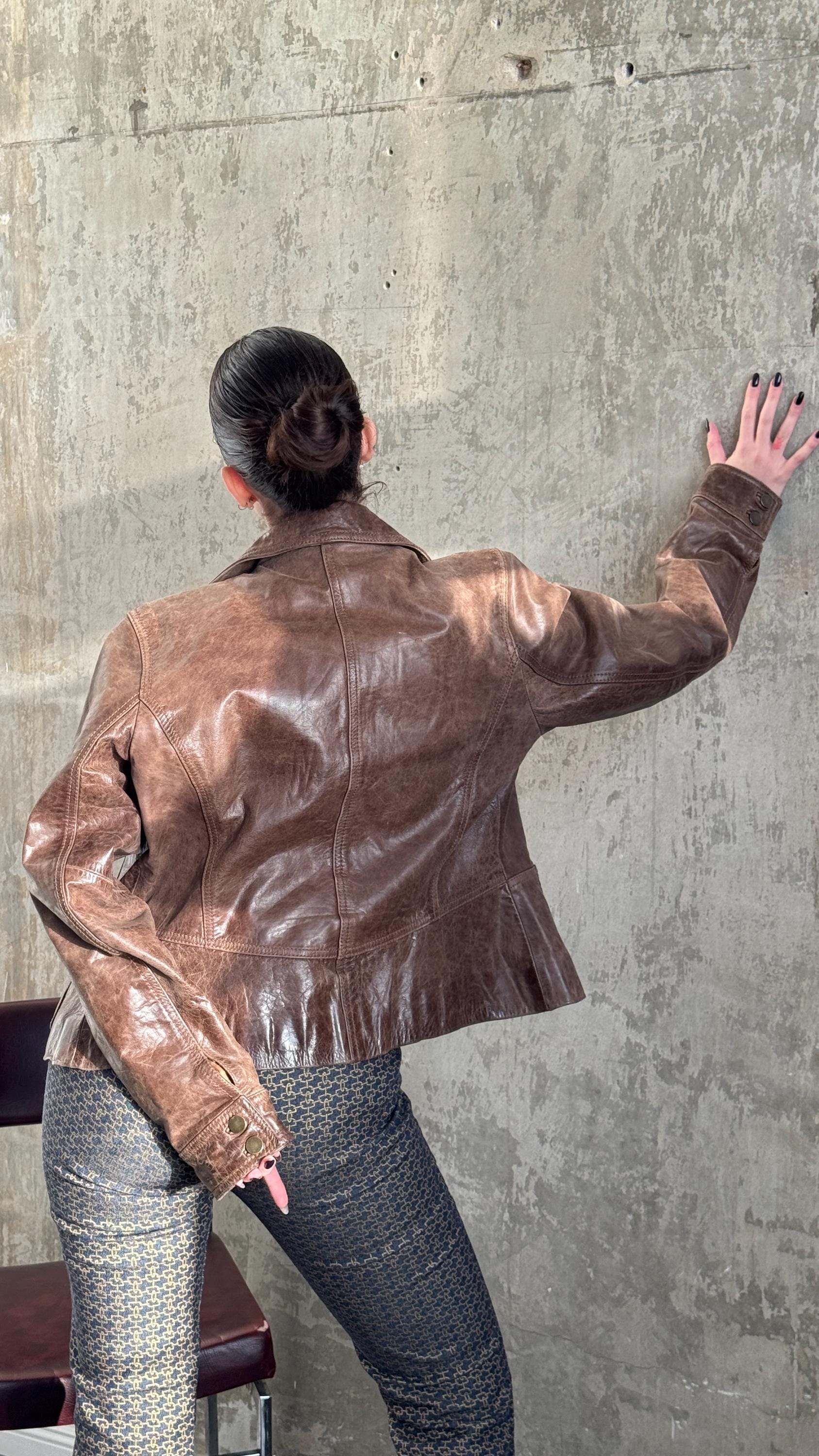 A person wearing a brown leather jacket stands in front of a concrete wall, with their back to the camera.