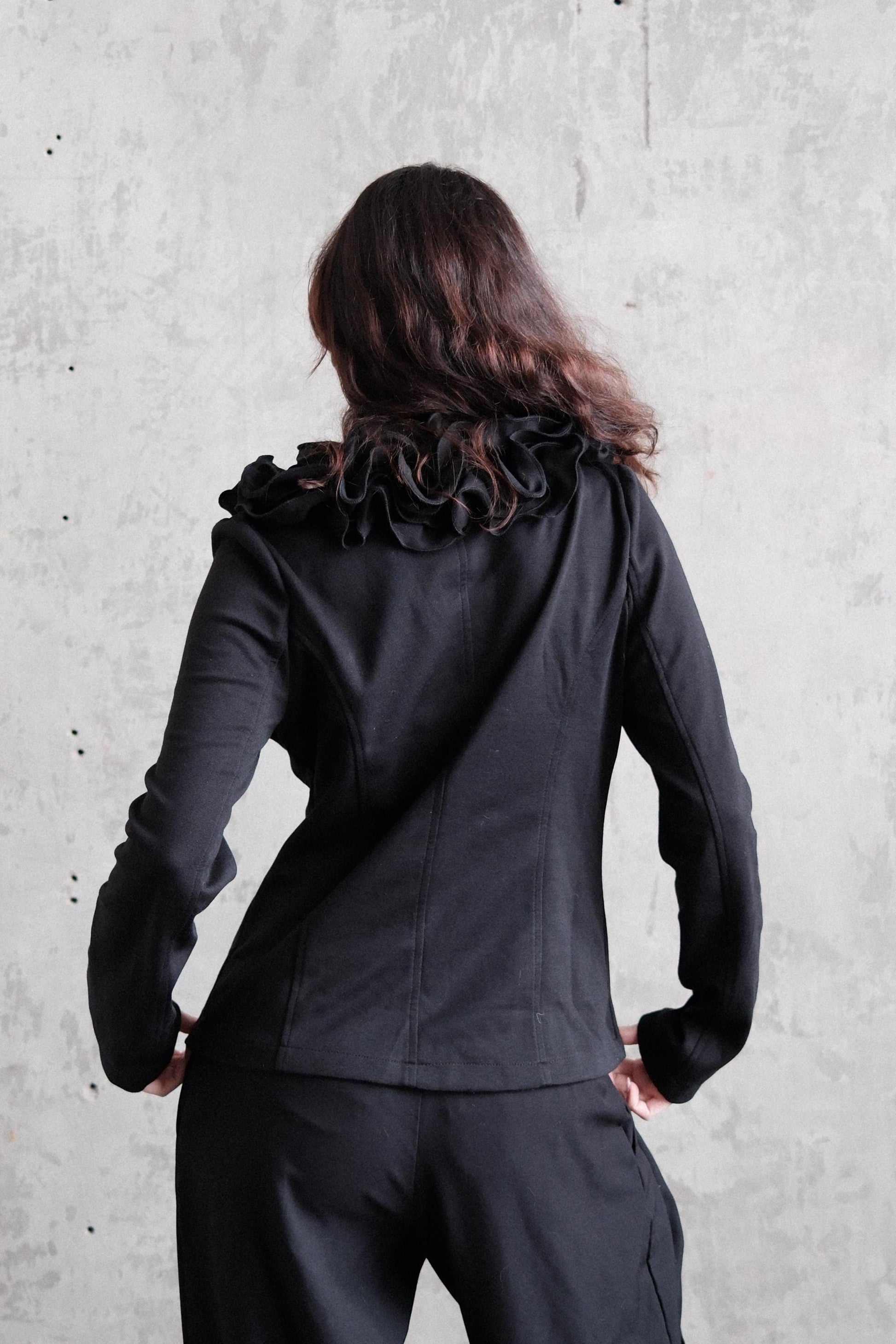 A person wearing a black jacket with a ruffled collar is standing against a plain concrete wall.