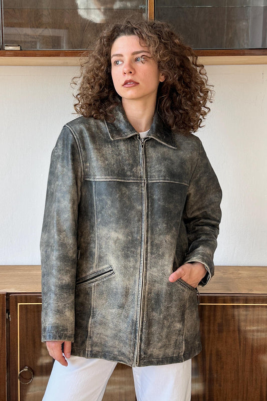 A woman with curly hair wearing a gray leather jacket stands in front of a wooden cabinet.