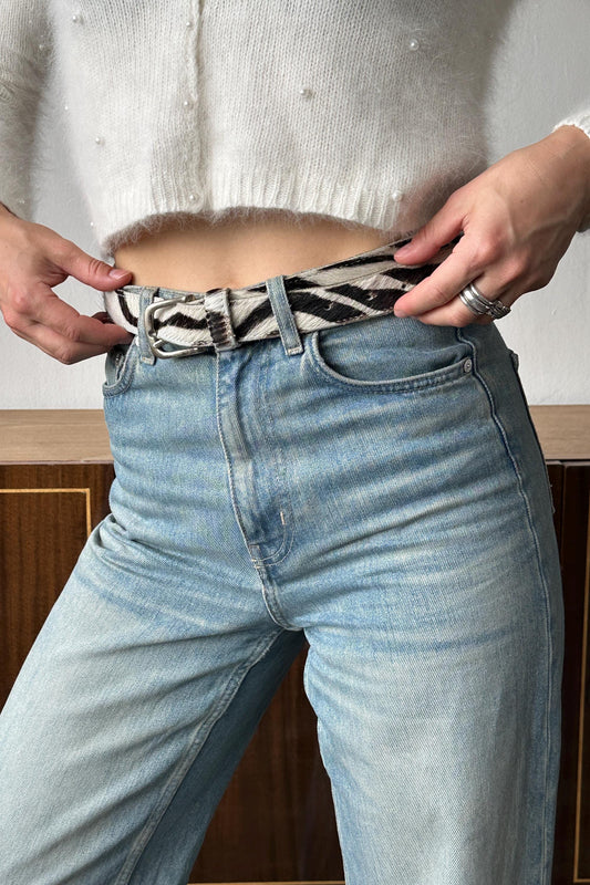 A person wearing a white sweater and blue jeans, with their hands adjusting a zebra-striped belt.