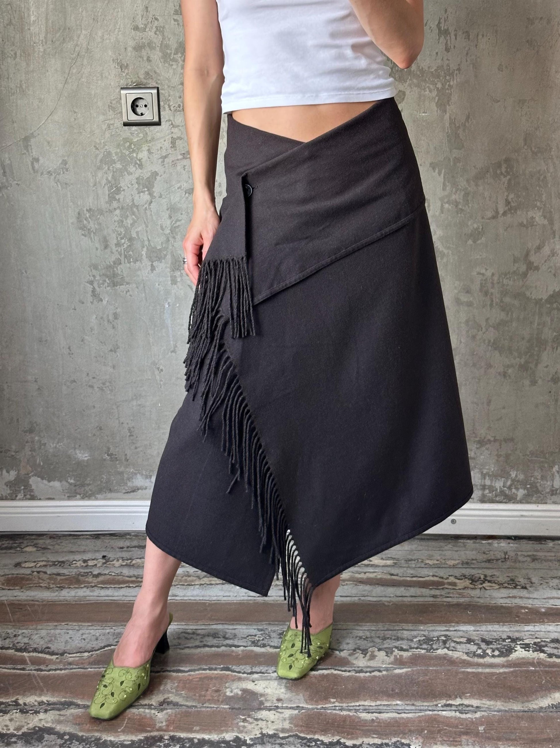 A woman wearing a black skirt with fringe details and green shoes stands in front of a concrete wall.