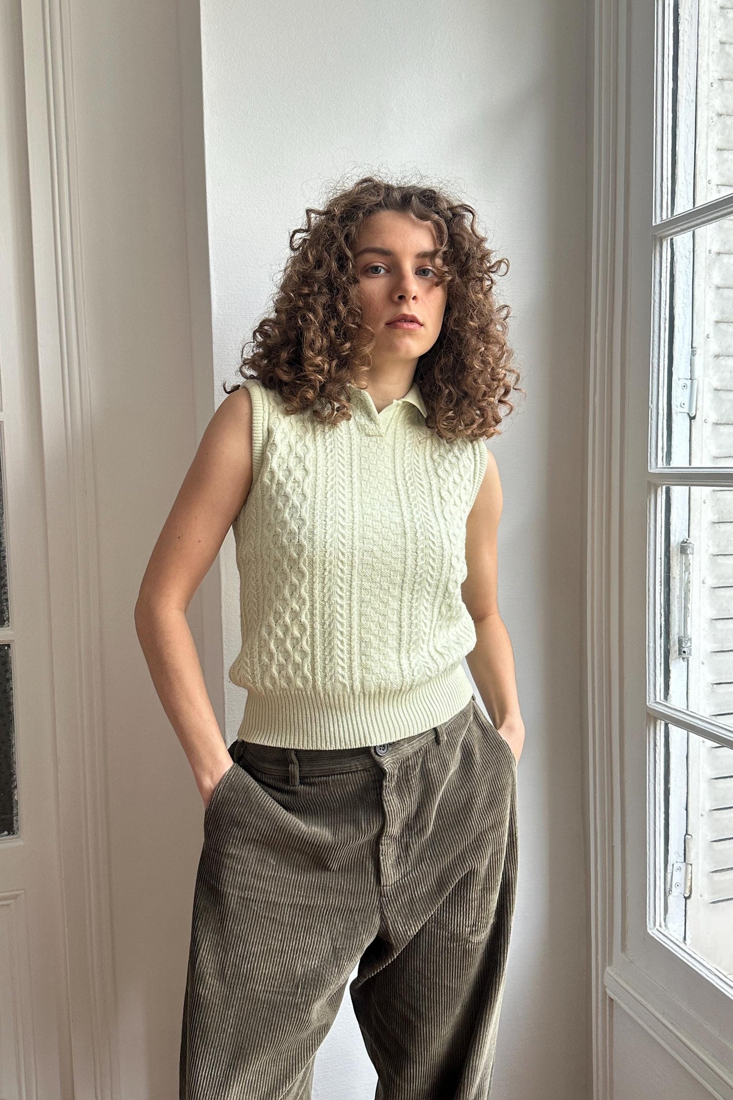 A woman with curly hair stands in front of a window, wearing a white sweater vest and gray pants.