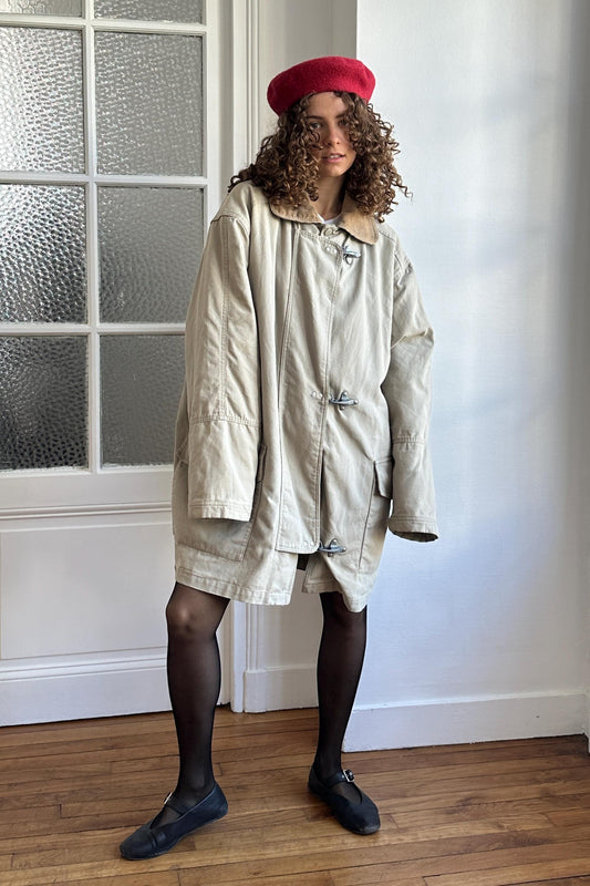 A person wearing a beige coat, black tights, and black shoes stands in front of a white door with a glass window.