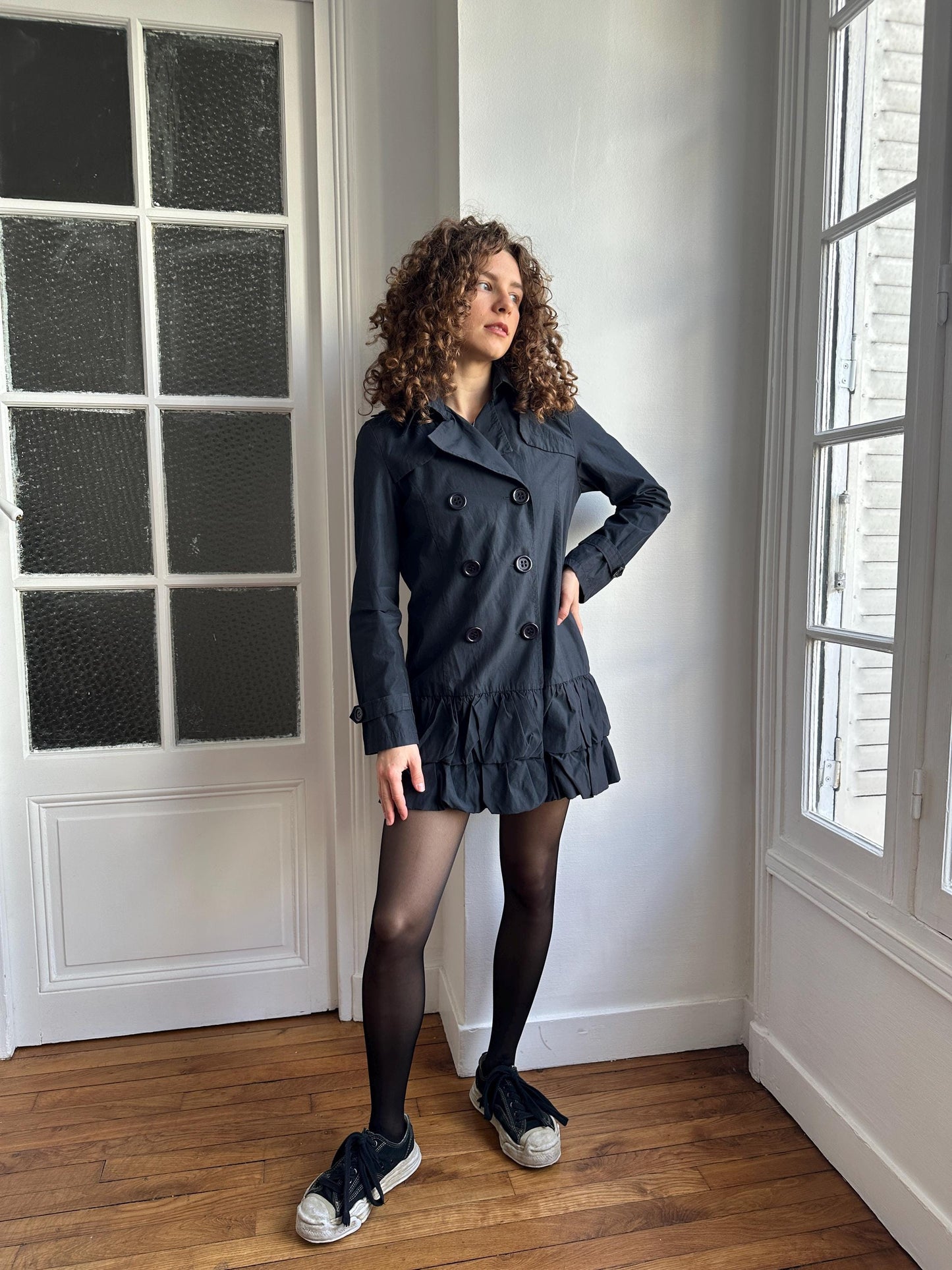 A woman with curly hair is standing in a room, wearing a black coat and black shoes.