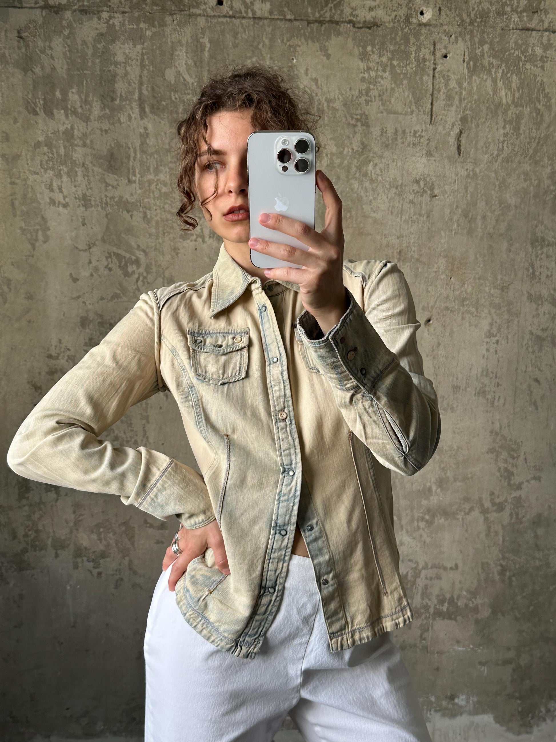 A person is taking a selfie with a smartphone, wearing a beige denim jacket and white pants, against a concrete wall background.