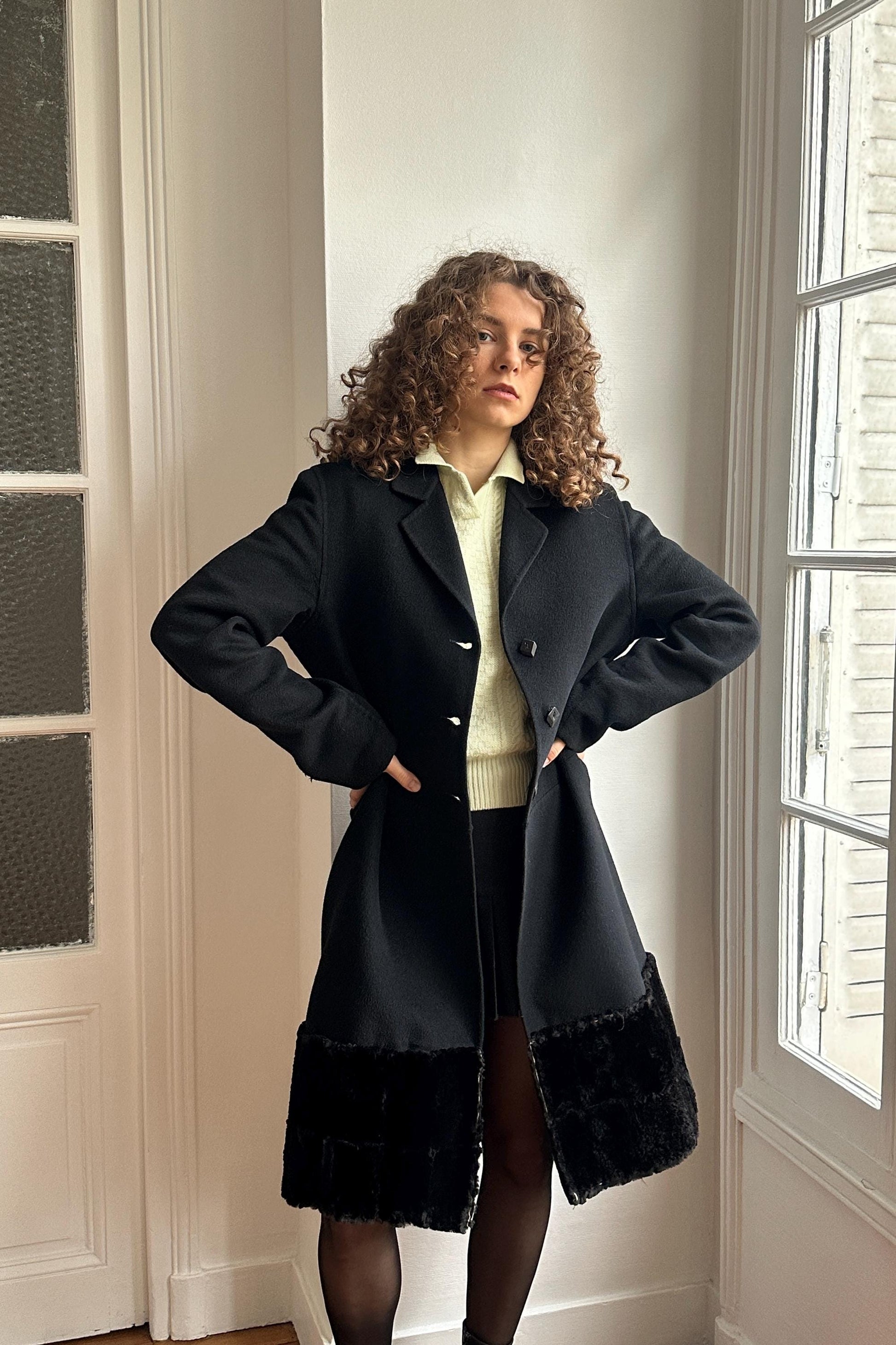 A woman with curly hair stands in a room, wearing a black coat with fur trim and posing with her hands on her hips.
