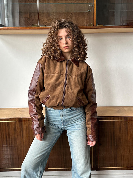 A person with curly hair wearing a brown jacket and blue jeans is standing in front of a wooden cabinet.