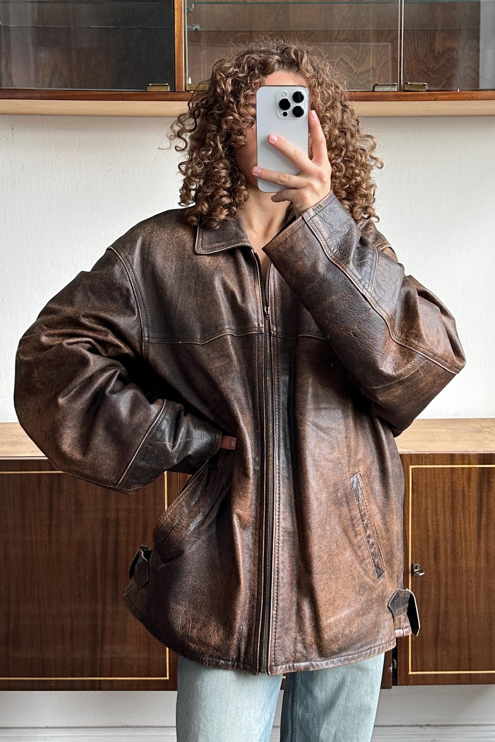 A person with curly hair is taking a selfie with a smartphone, wearing a brown leather jacket.