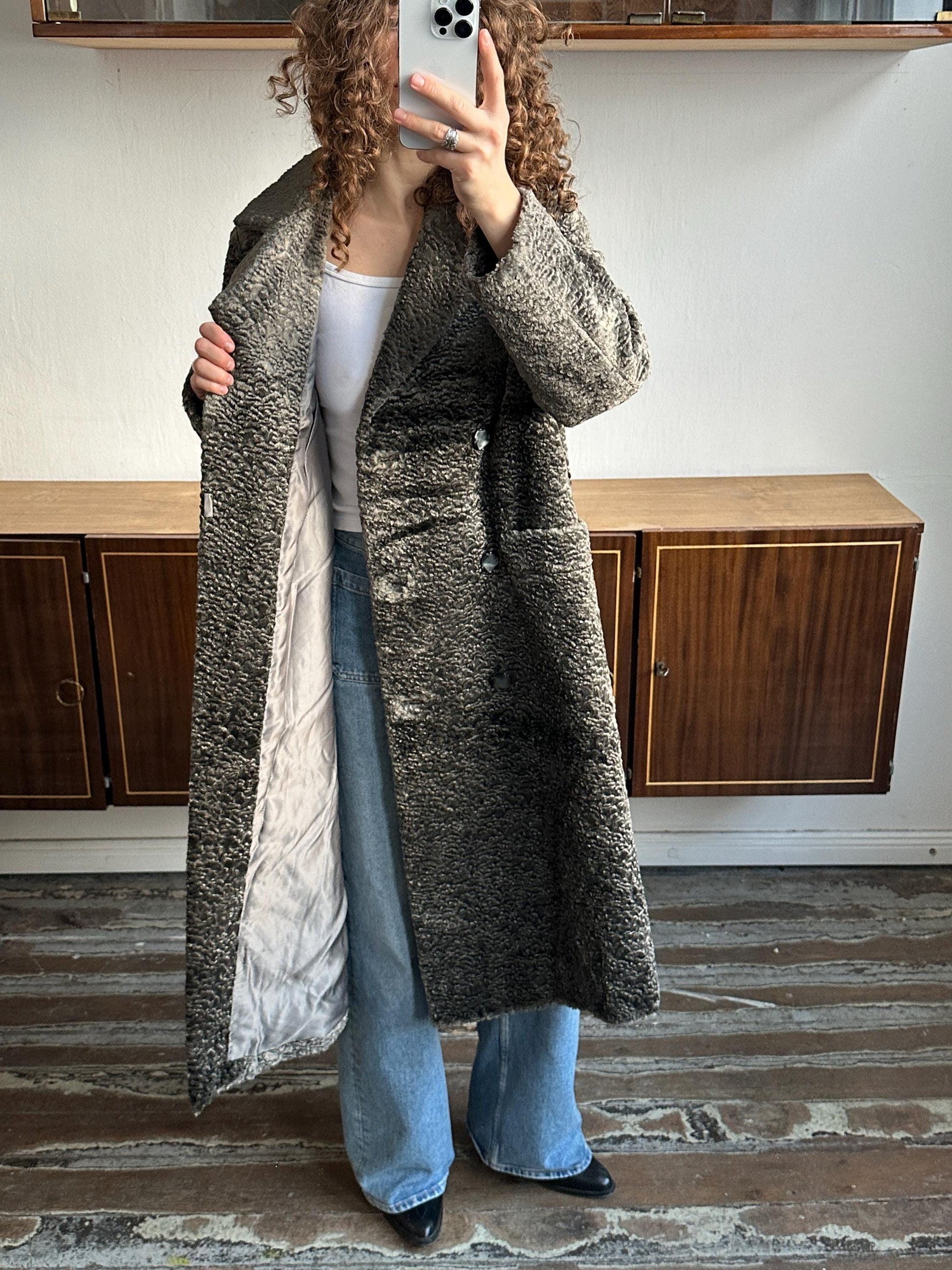 A person is taking a selfie with a phone, wearing a long gray coat and blue jeans.