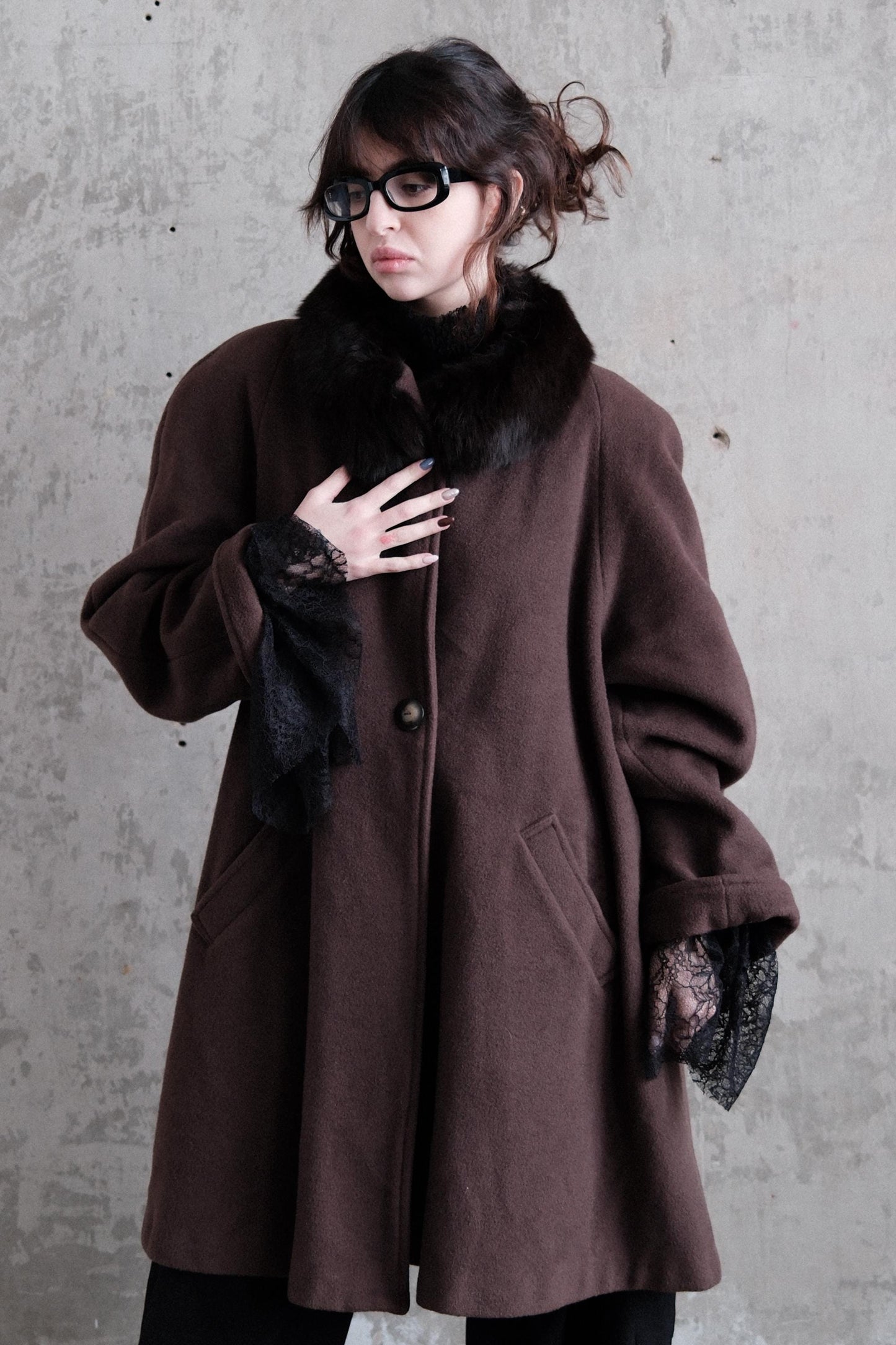 A woman wearing a brown coat with lace sleeves and glasses is standing against a concrete wall.