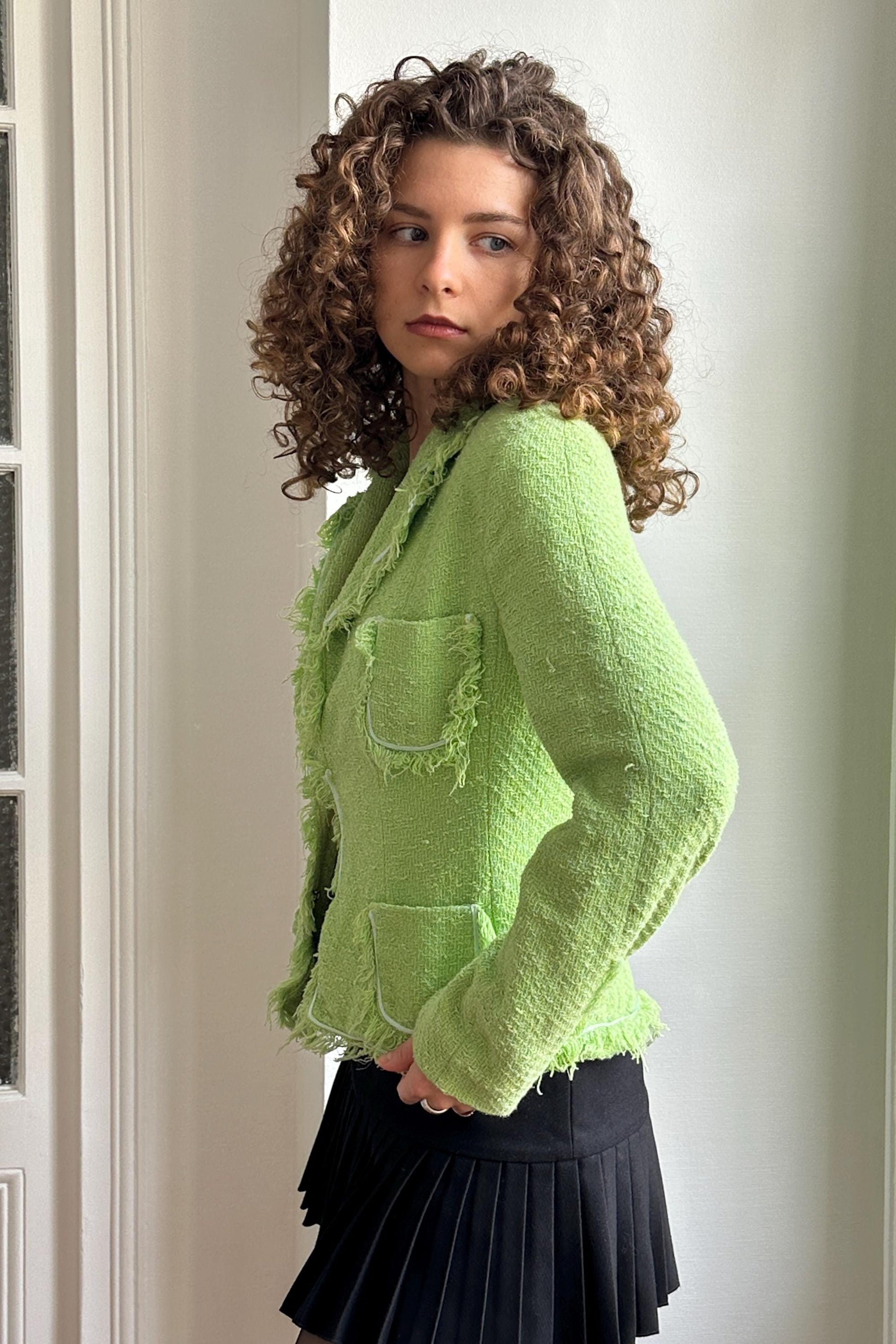 A young woman with curly hair wearing a lime green tweed jacket and a black skirt stands in a room.