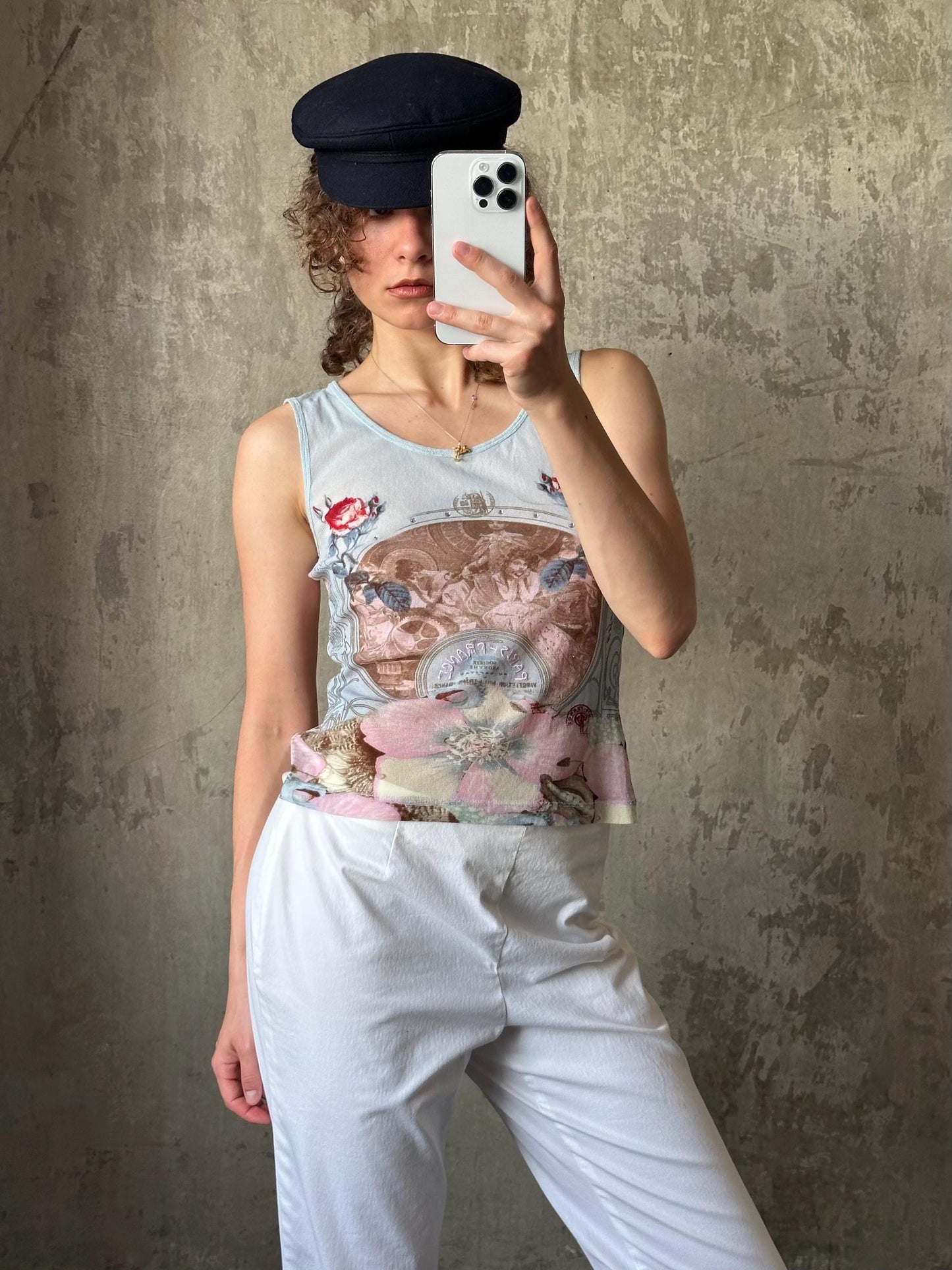 A woman is taking a selfie with a smartphone, wearing a white tank top with a floral design and white pants.