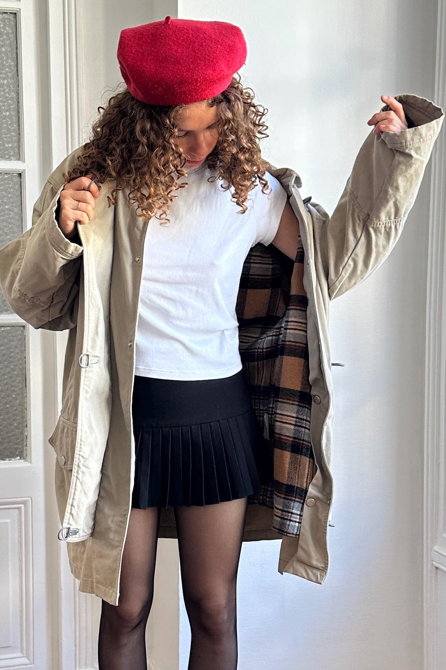 A woman with curly hair wearing a red beret, a beige coat, and a black pleated skirt stands in a room, her arms outstretched.