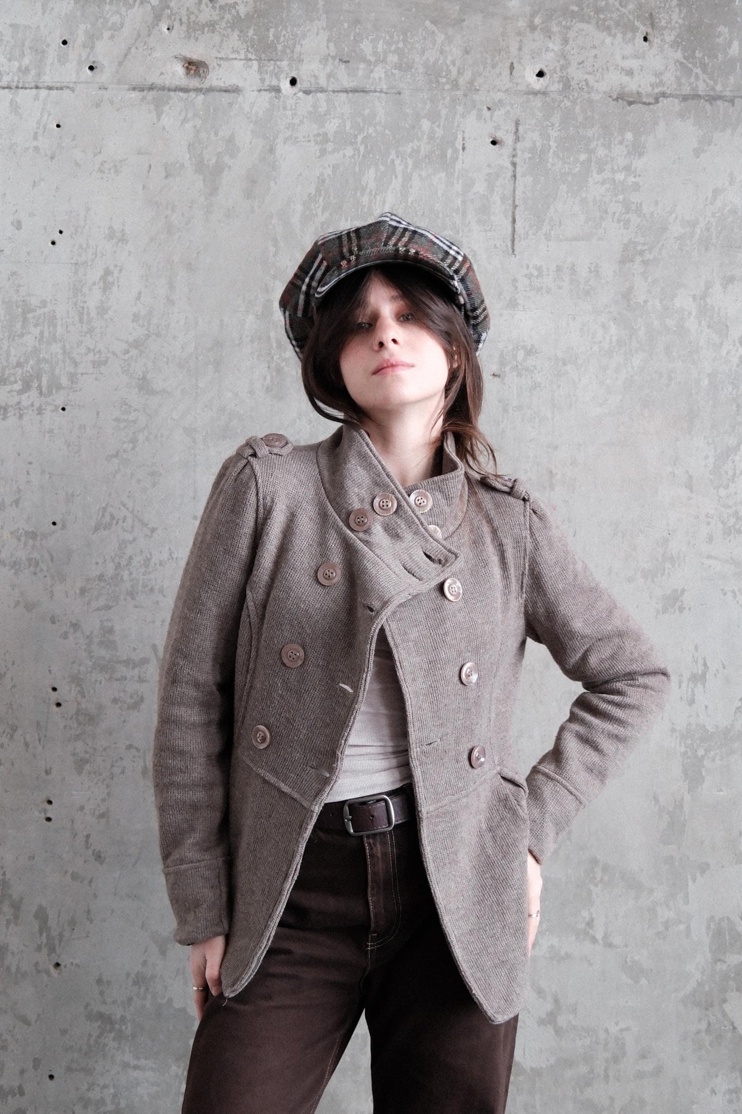 A woman wearing a gray coat and a plaid hat stands against a concrete wall, her gaze directed off to the side.