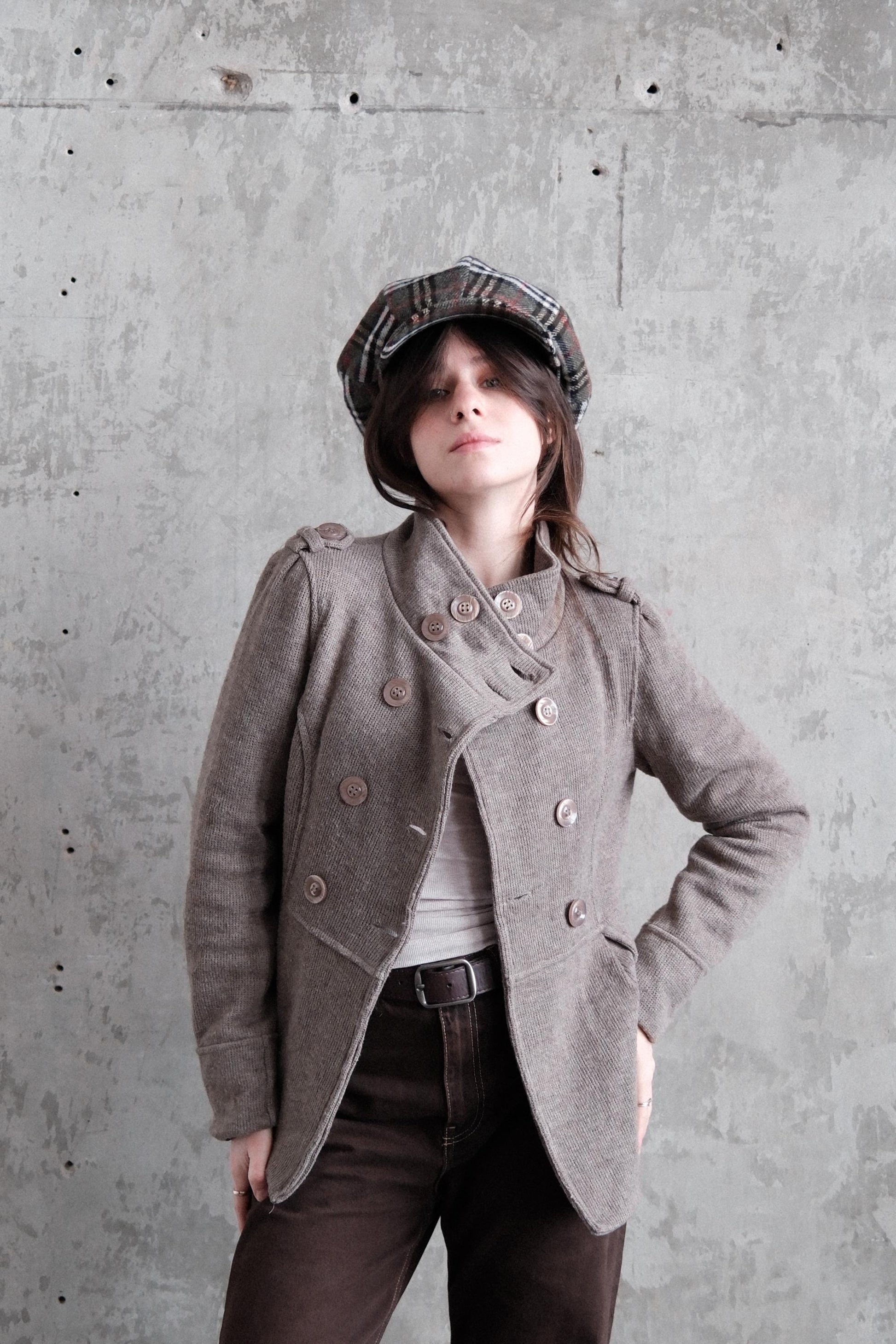 A woman wearing a gray coat and a plaid hat stands against a concrete wall, her gaze directed off to the side.