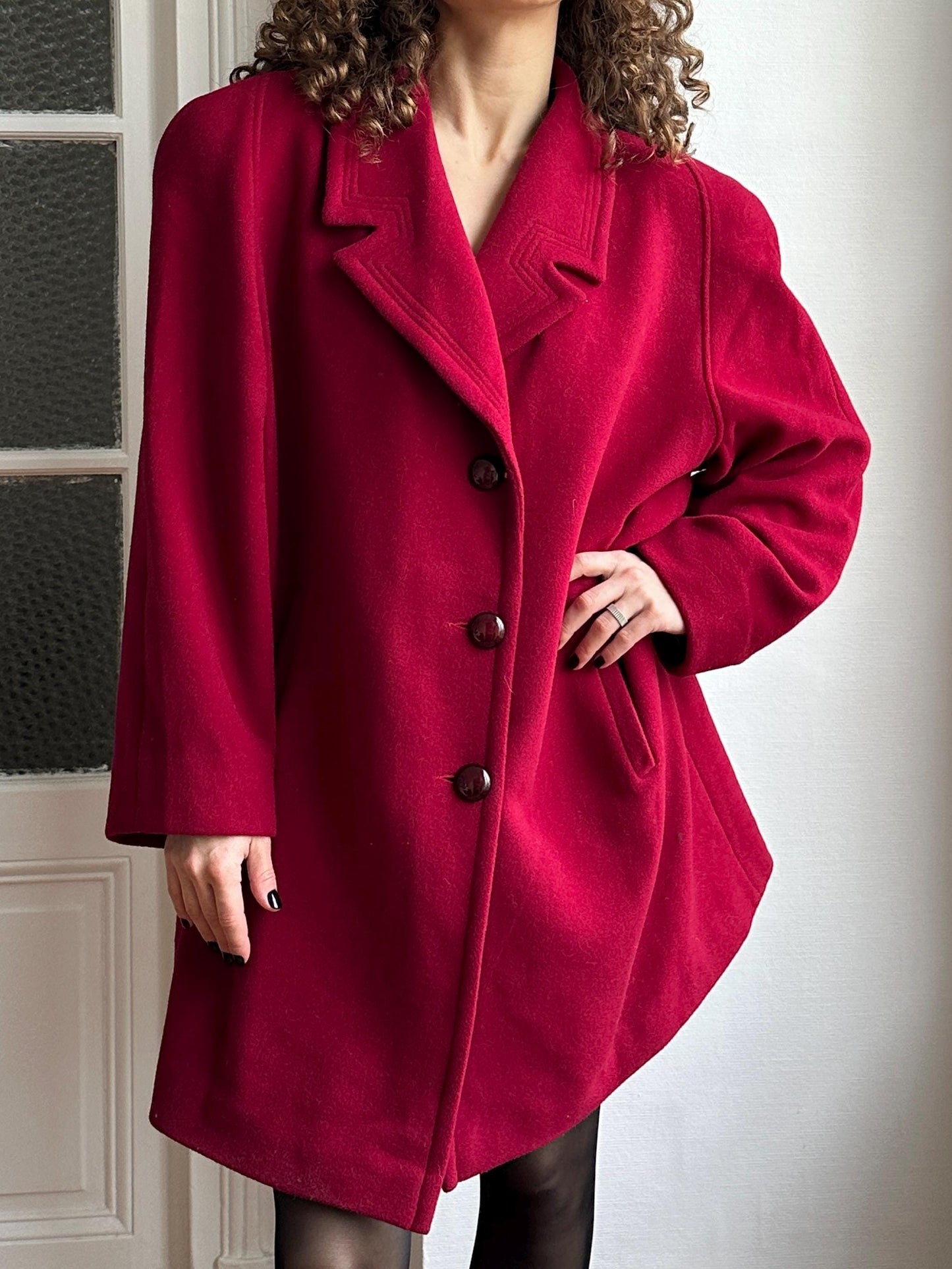 A woman wearing a red coat with a high collar and three buttons, standing in front of a white door.