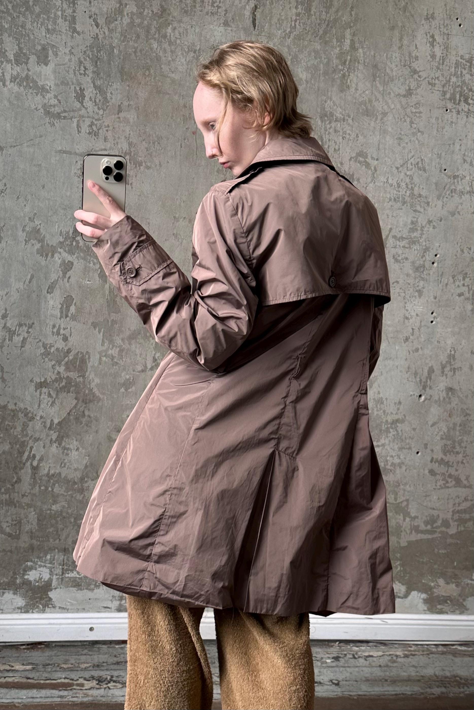A person wearing a brown coat is standing in front of a concrete wall, holding a phone and looking at it.