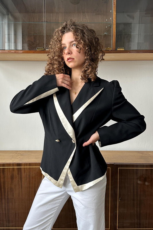 A woman with curly hair wearing a black and white blazer stands in front of a wooden cabinet.