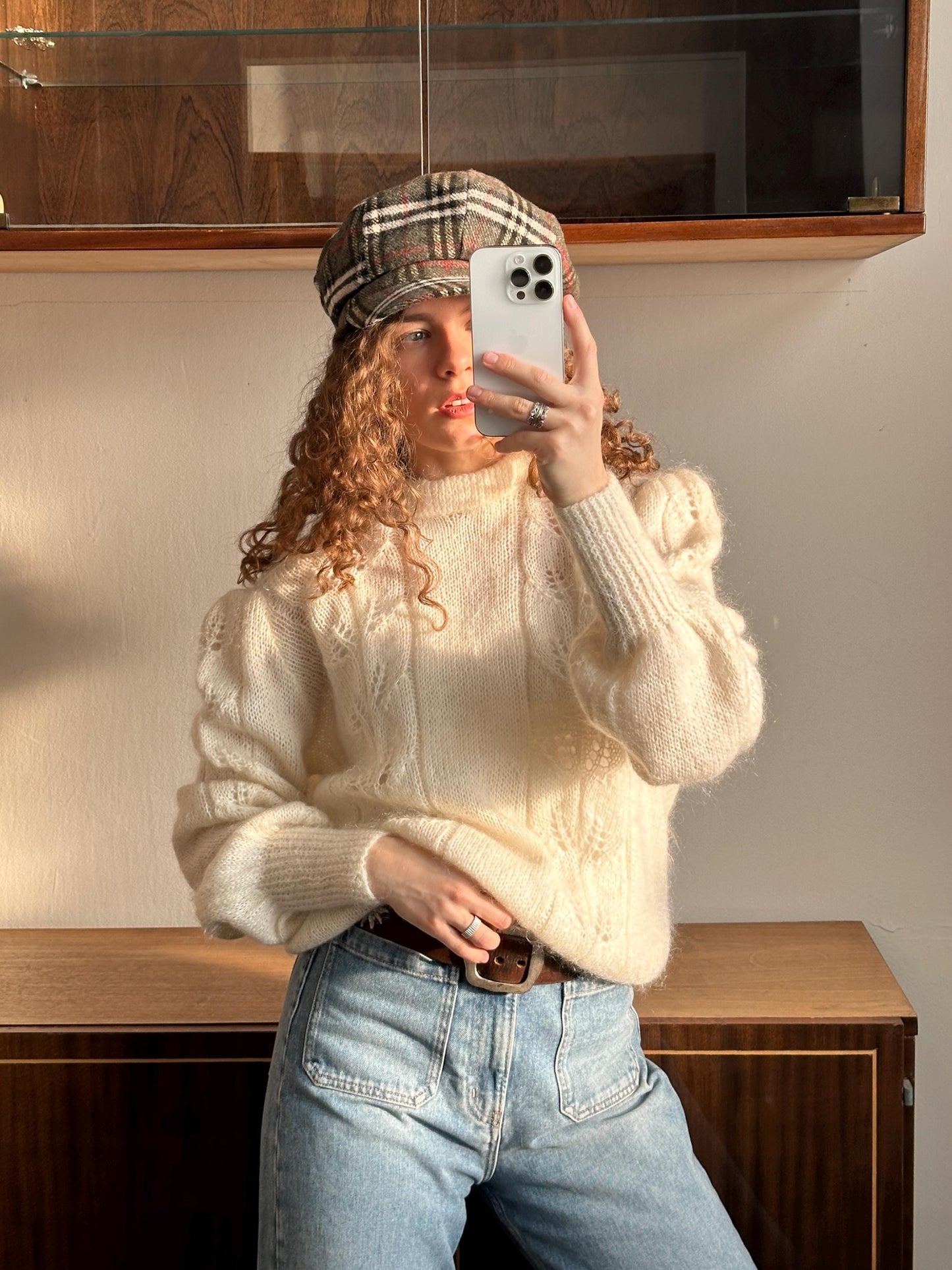 A woman with curly hair is taking a selfie with a white iPhone, wearing a plaid hat and a white sweater.