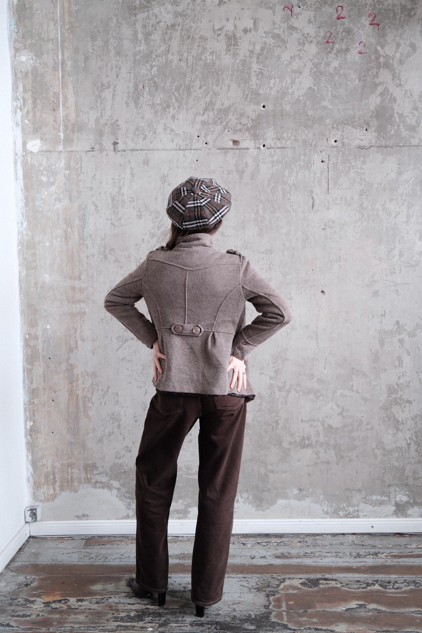 a person standing in a room with a concrete wall and a wooden floor. The person is wearing a brown jacket and a plaid hat.