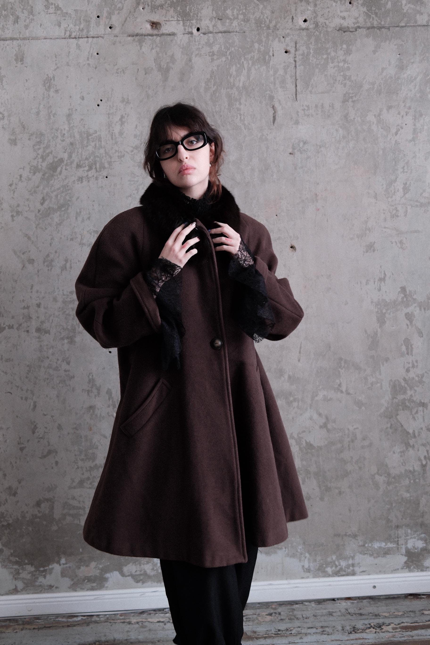 A woman wearing a brown coat and glasses stands against a concrete wall, looking to the side.