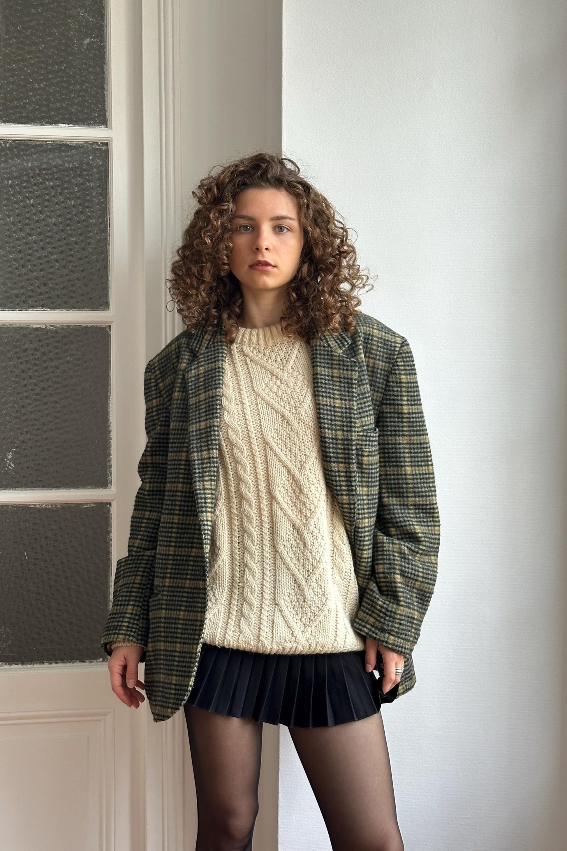 A young woman with curly hair stands in front of a white door, wearing a plaid jacket, a white sweater, and a black skirt.