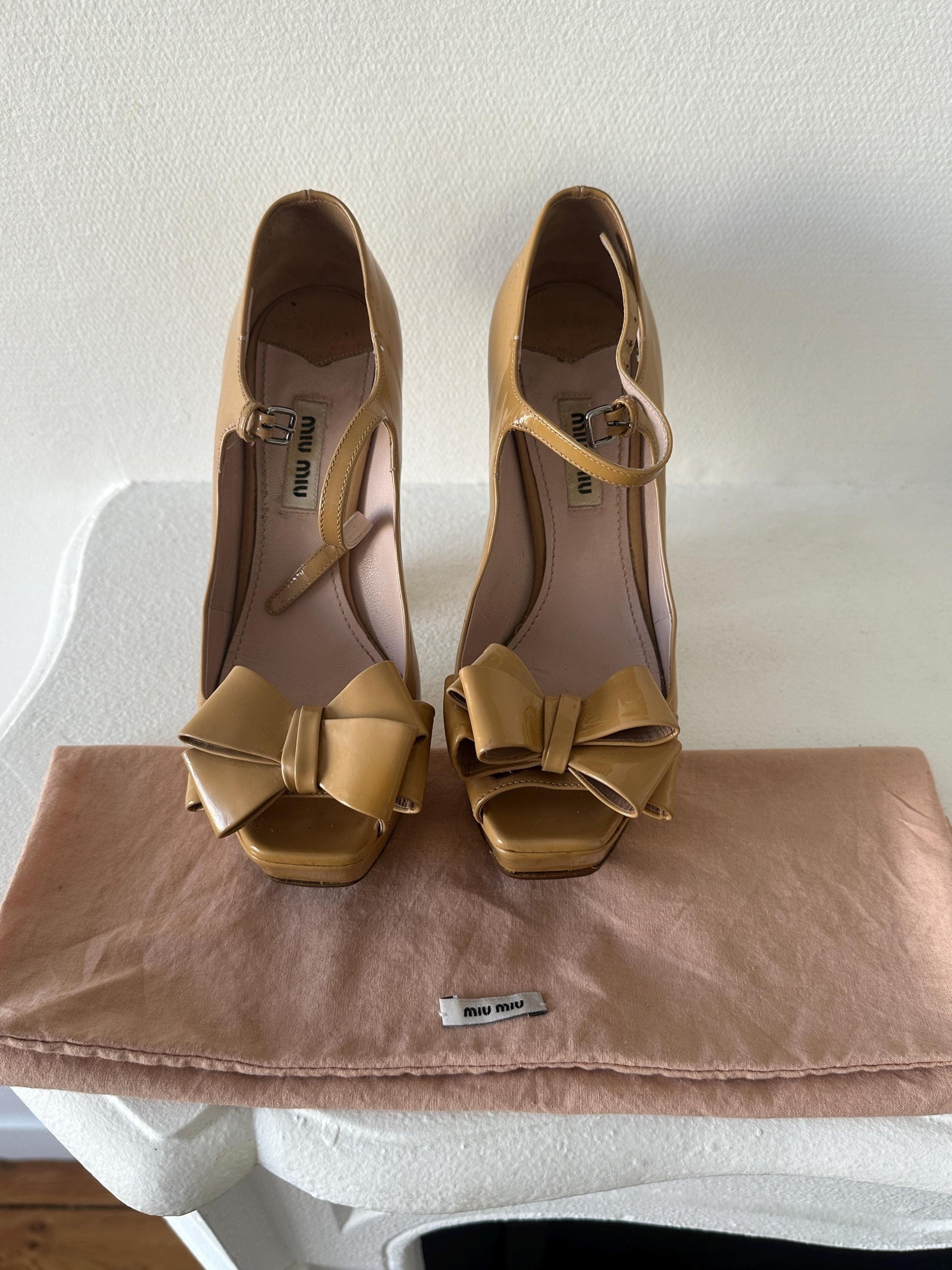 A pair of beige high-heeled shoes with bows on the toe and heel, placed on a white surface next to a brown satin bag.