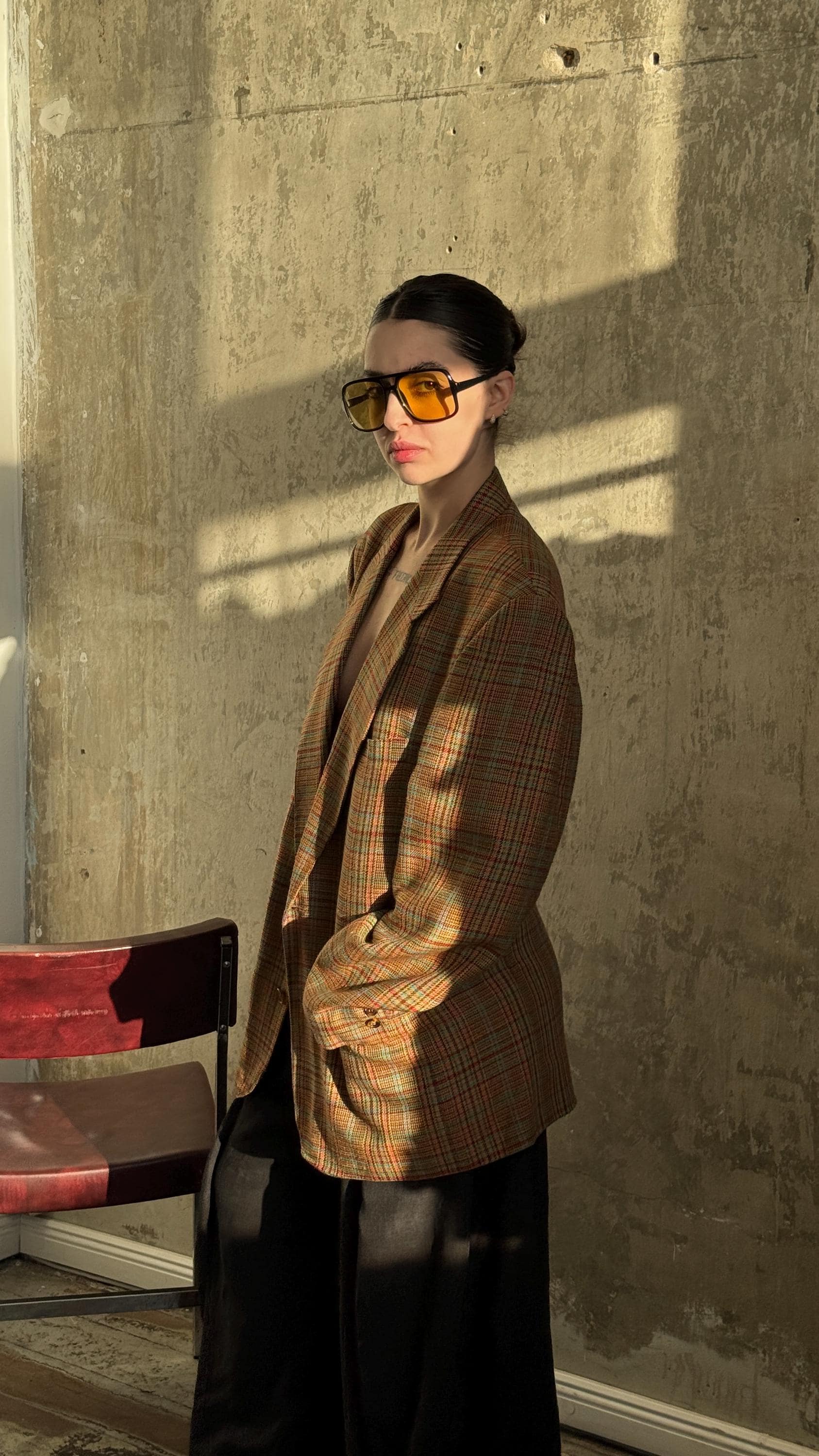 A woman wearing a plaid jacket and sunglasses stands in front of a concrete wall, with a red chair visible to her side.