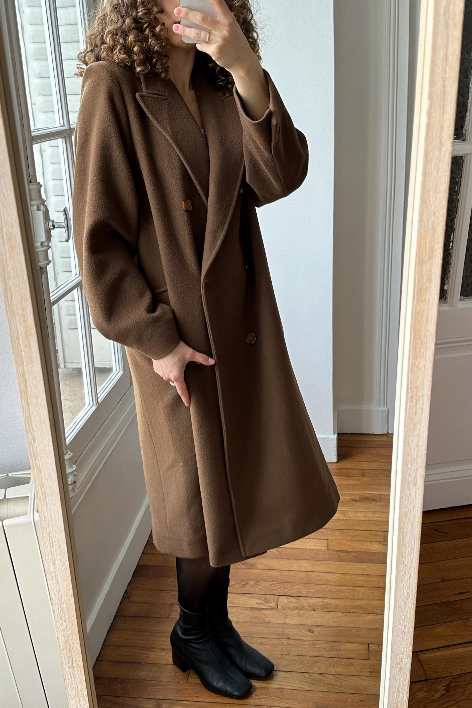 A woman wearing a brown coat is taking a mirror selfie in a room with wooden floors.